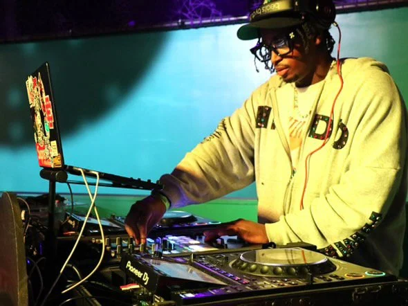 DJ wearing a beige hoodie and glasses, standing behind DJ equipment with a colorful background.