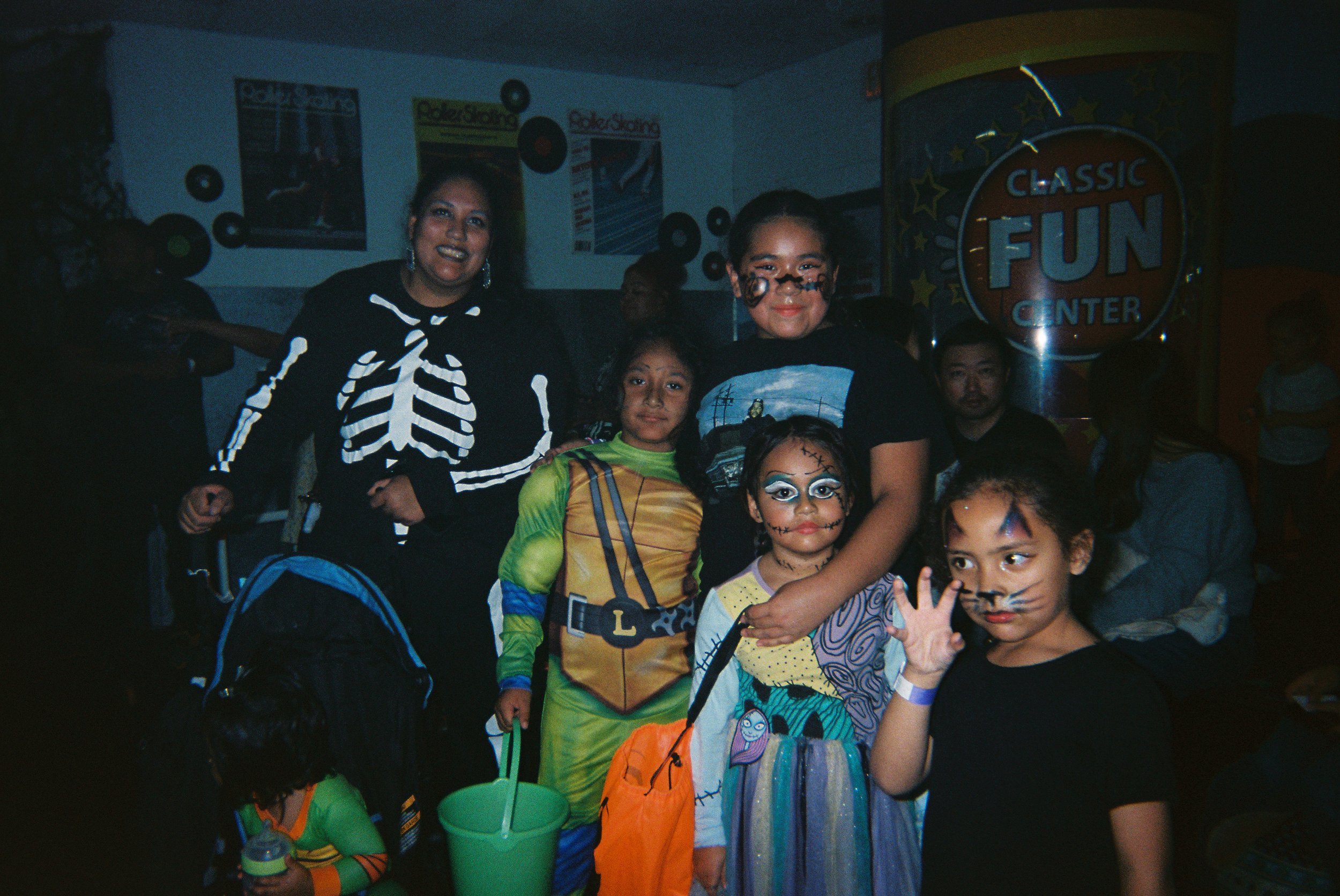 Children and adults in costumes at a Halloween party, some with face paint, in an indoor fun center with a sign that reads 'Classic Fun Center'.