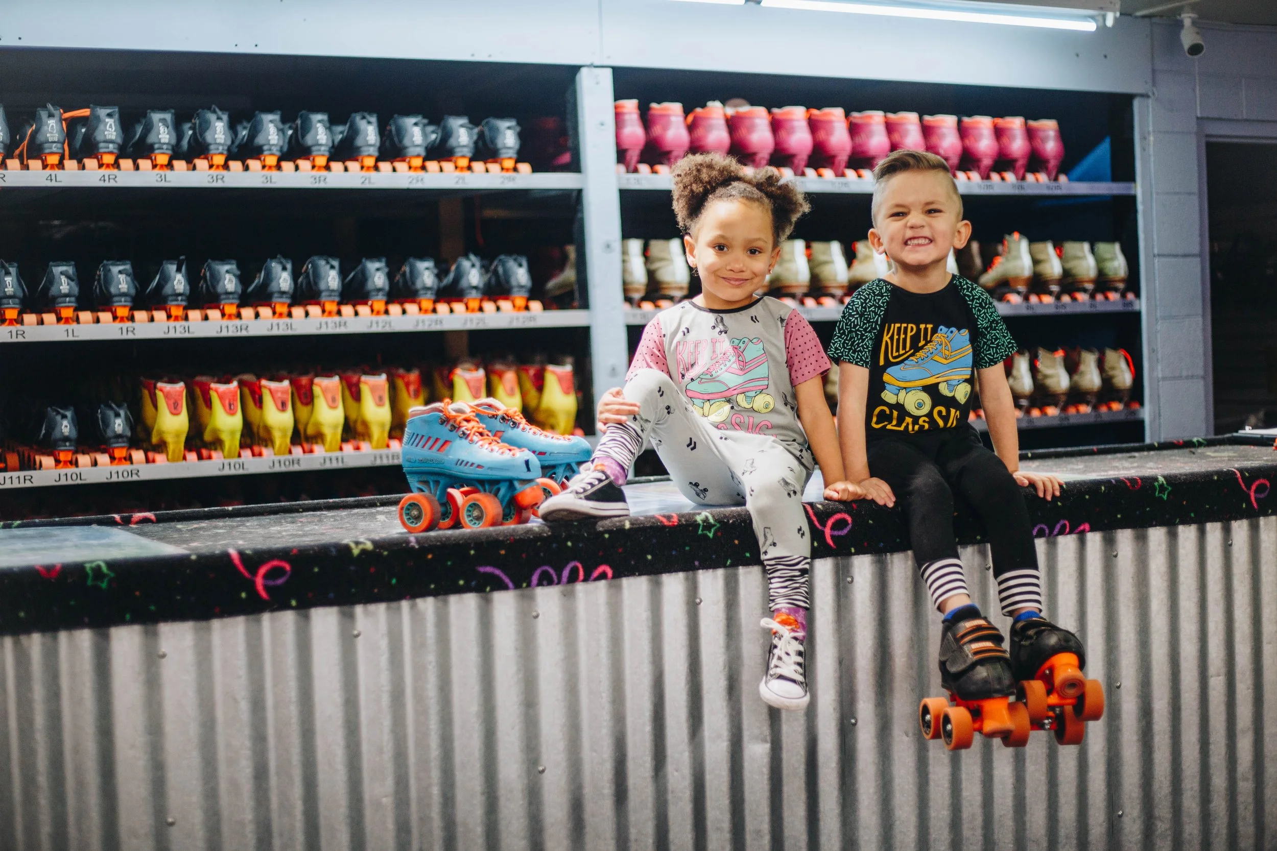 Two children sitting on the edge of a skating rink with roller skates on their feet, smiling at the camera, with shelves of colorful roller skates in the background.