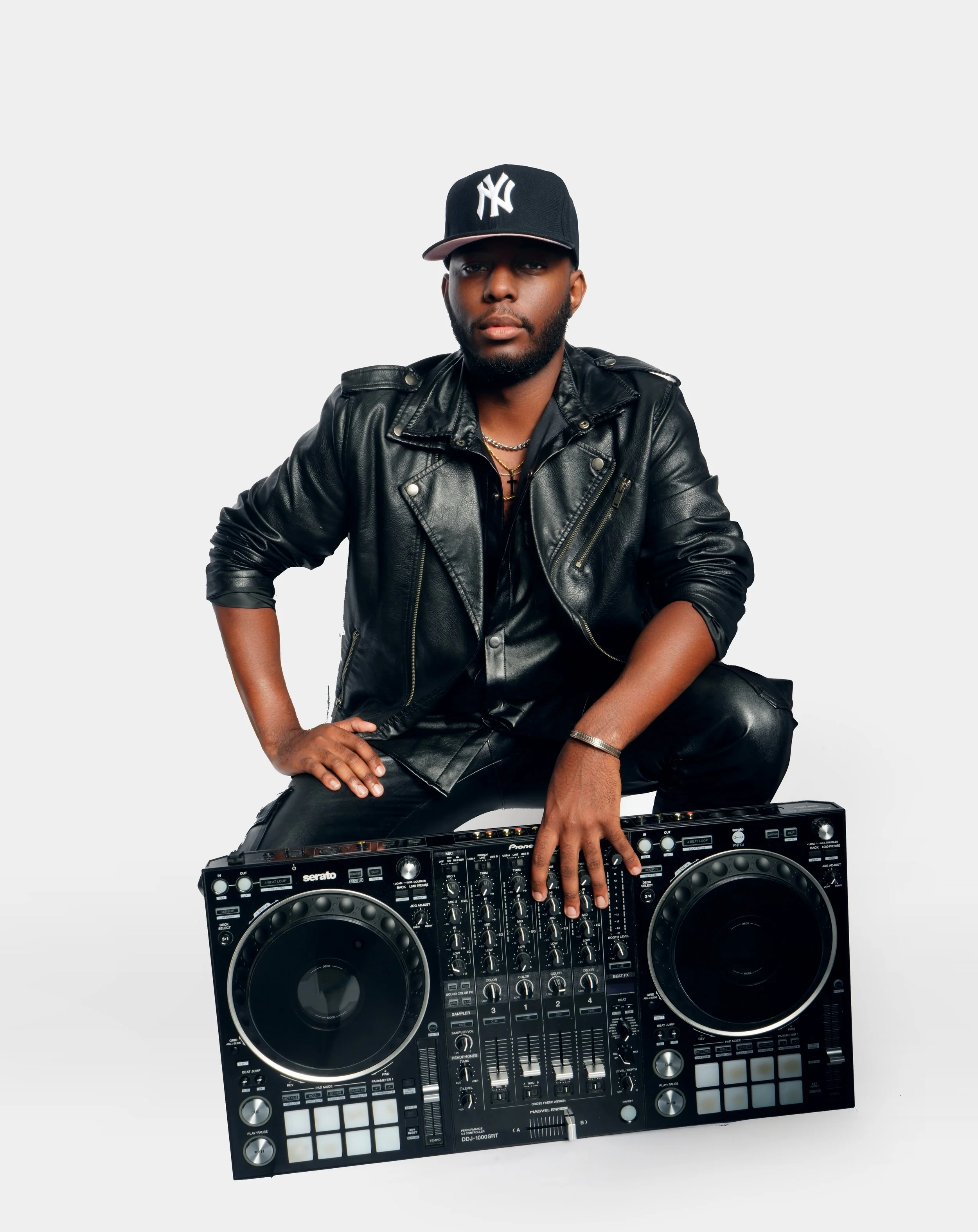 A DJ wearing a black leather jacket, black cap, and jewelry, crouching behind a DJ controller.