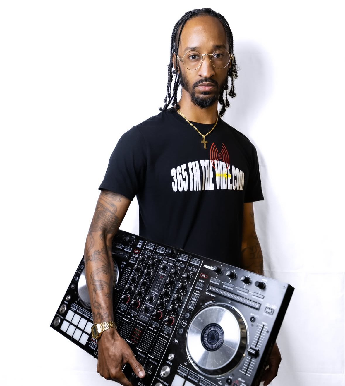 A man with long braided hair, glasses, and tattoos on his arms is holding a DJ controller. He is wearing a black T-shirt with the website 365entertainment.com printed on it, a gold watch, and a gold cross necklace, standing against a plain white back