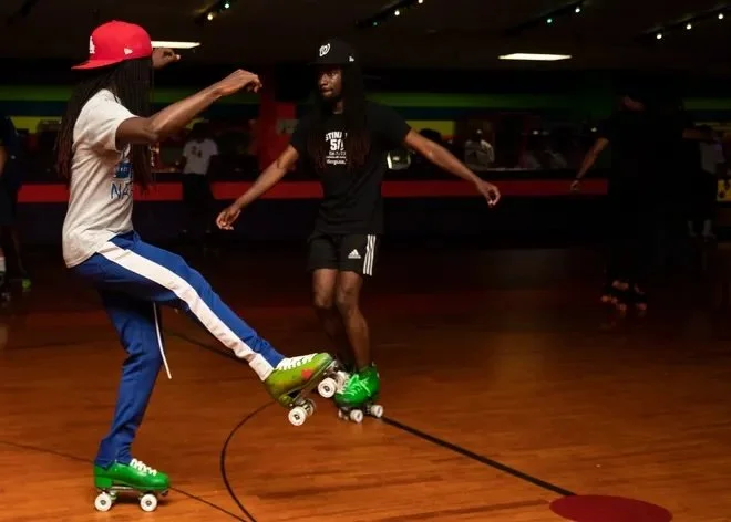 Two young men roller skating indoors, one wearing a red cap and the other a black cap, with a colorful wall and other skaters in the background.