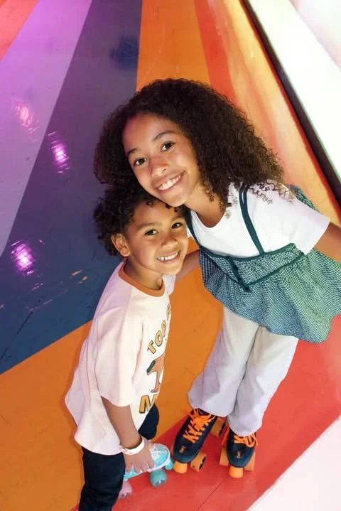 Two smiling children, a girl and a boy, standing on colorful roller skates in front of a vibrant rainbow-colored wall.