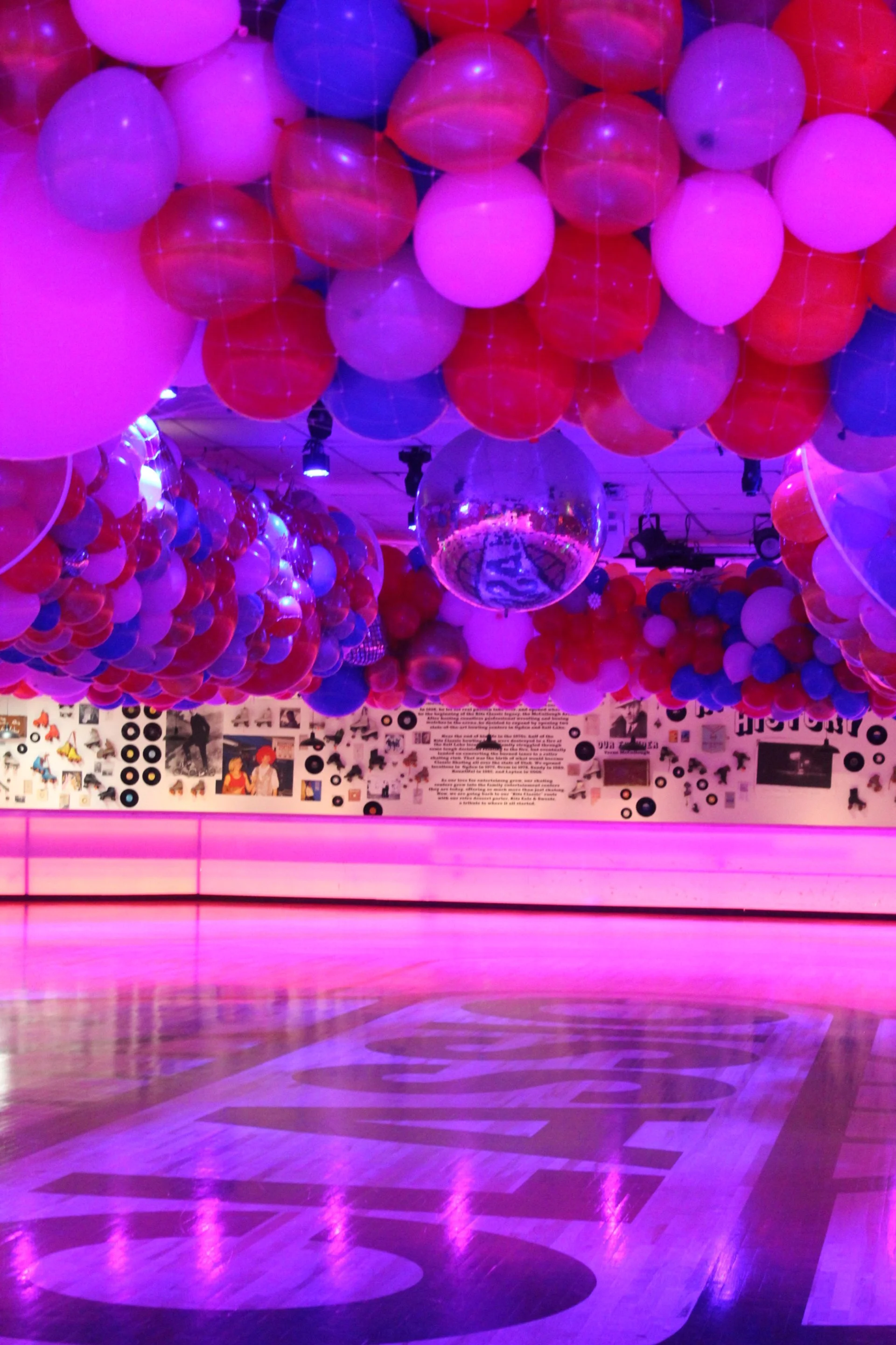 A dance floor with pink and purple lighting, decorated with numerous balloons in red, pink, purple, and blue, and a disco ball hanging from the ceiling.