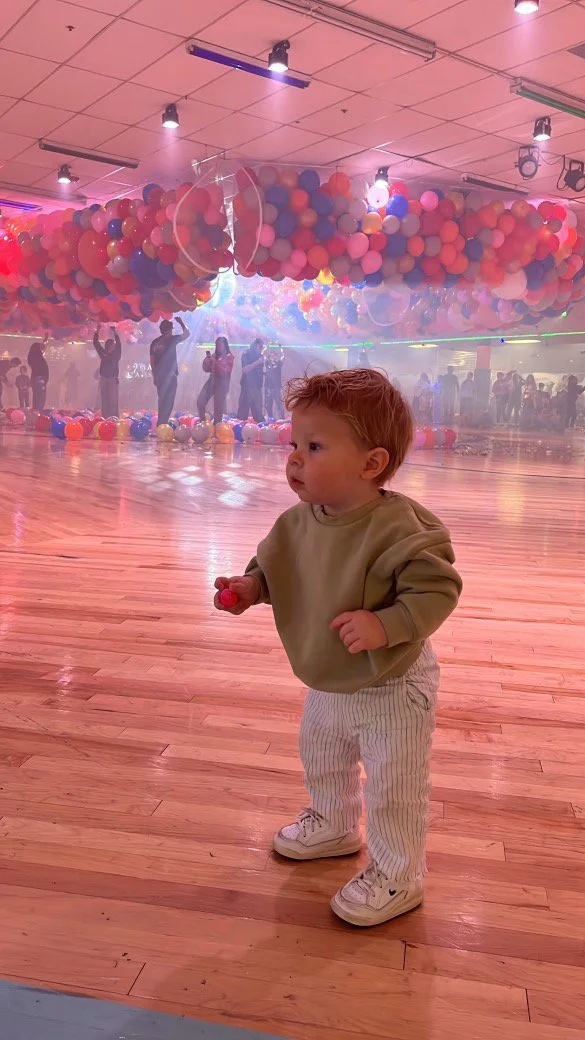 A young child with red hair, wearing a beige sweatshirt and striped pants, standing on a wooden floor at a ball pit or play area filled with many pink, red, and purple balloons, with people and balloon decorations in the background.