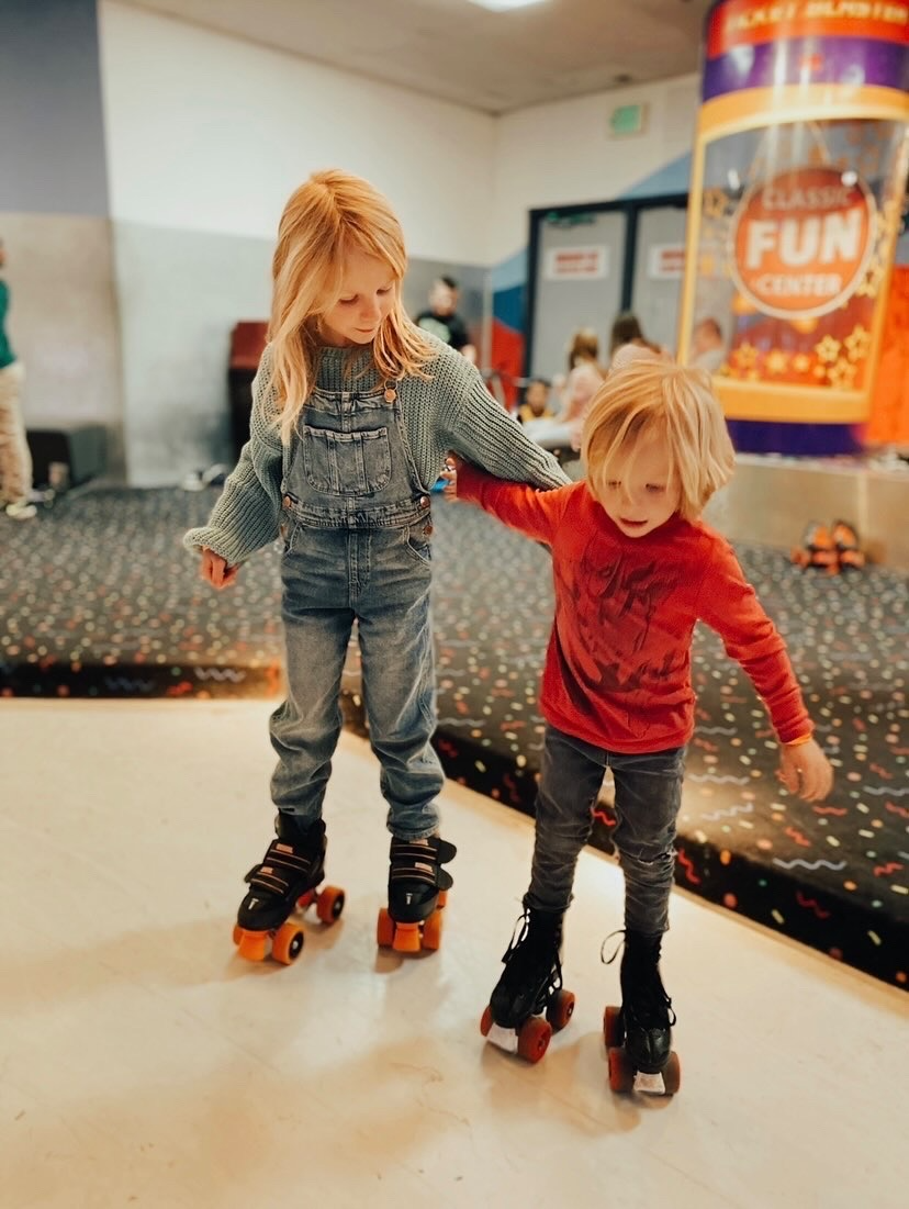 Two children roller skating indoors, with one helping the other with balance, in a recreational area with bunting and a sign in the background.