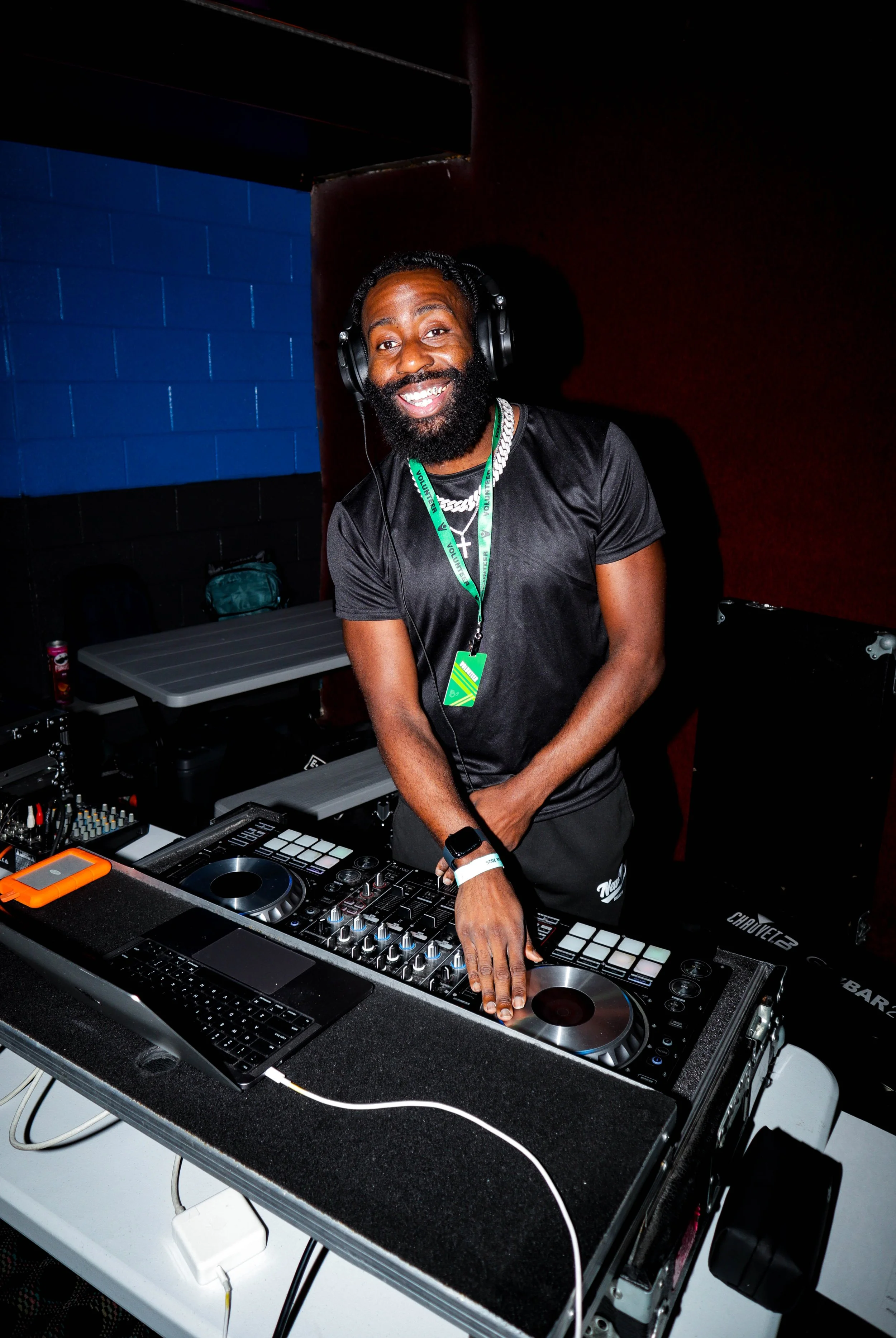 A smiling man with a beard and headphones standing behind a DJ booth, wearing a black shirt, indoors with a blue and red background.
