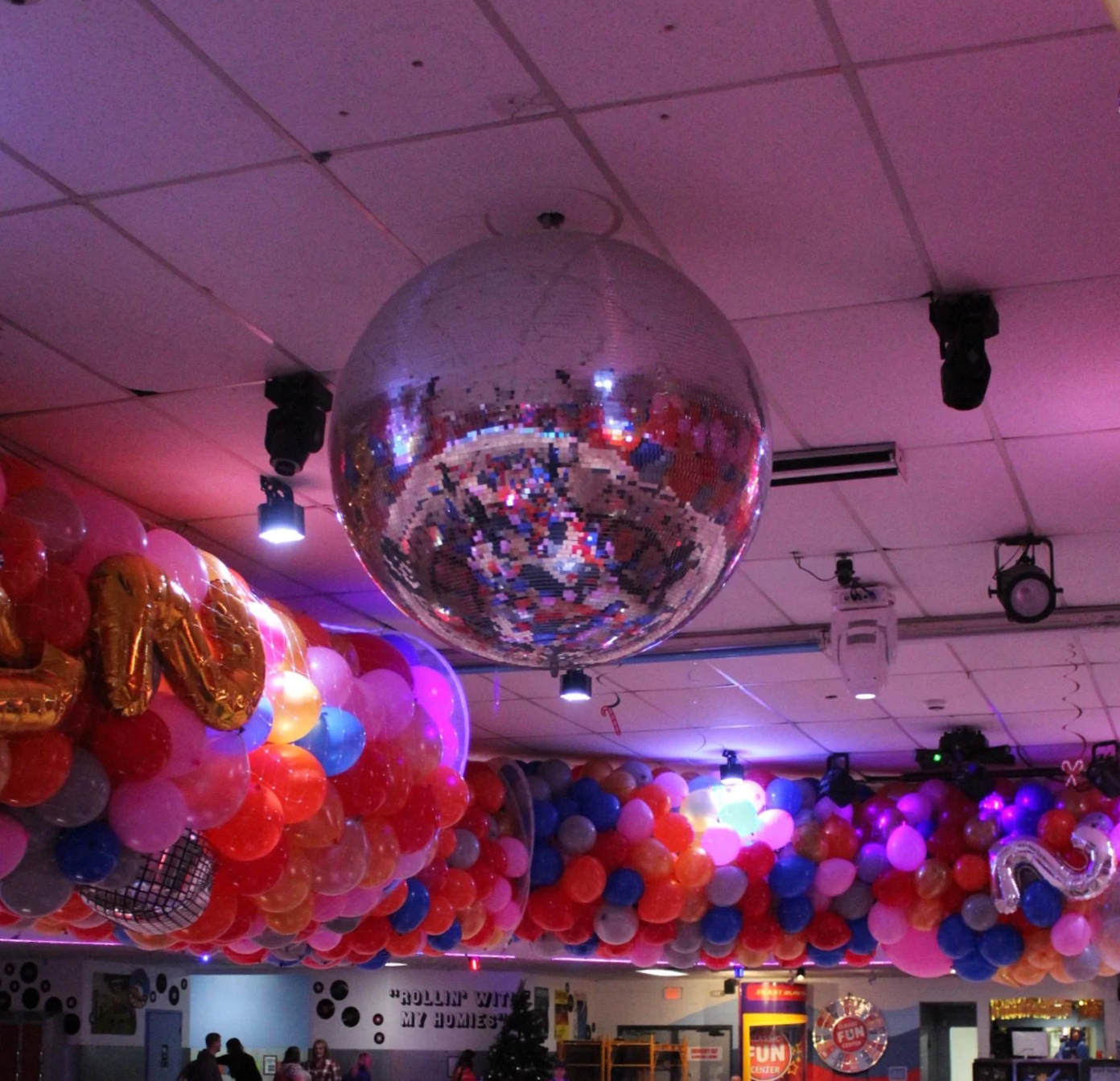 Decorated room with a large disco ball hanging from the ceiling, surrounded by colorful balloons including pink, red, blue, purple, and gold, with some balloons shaped like letters and numbers, and people visible in the background.