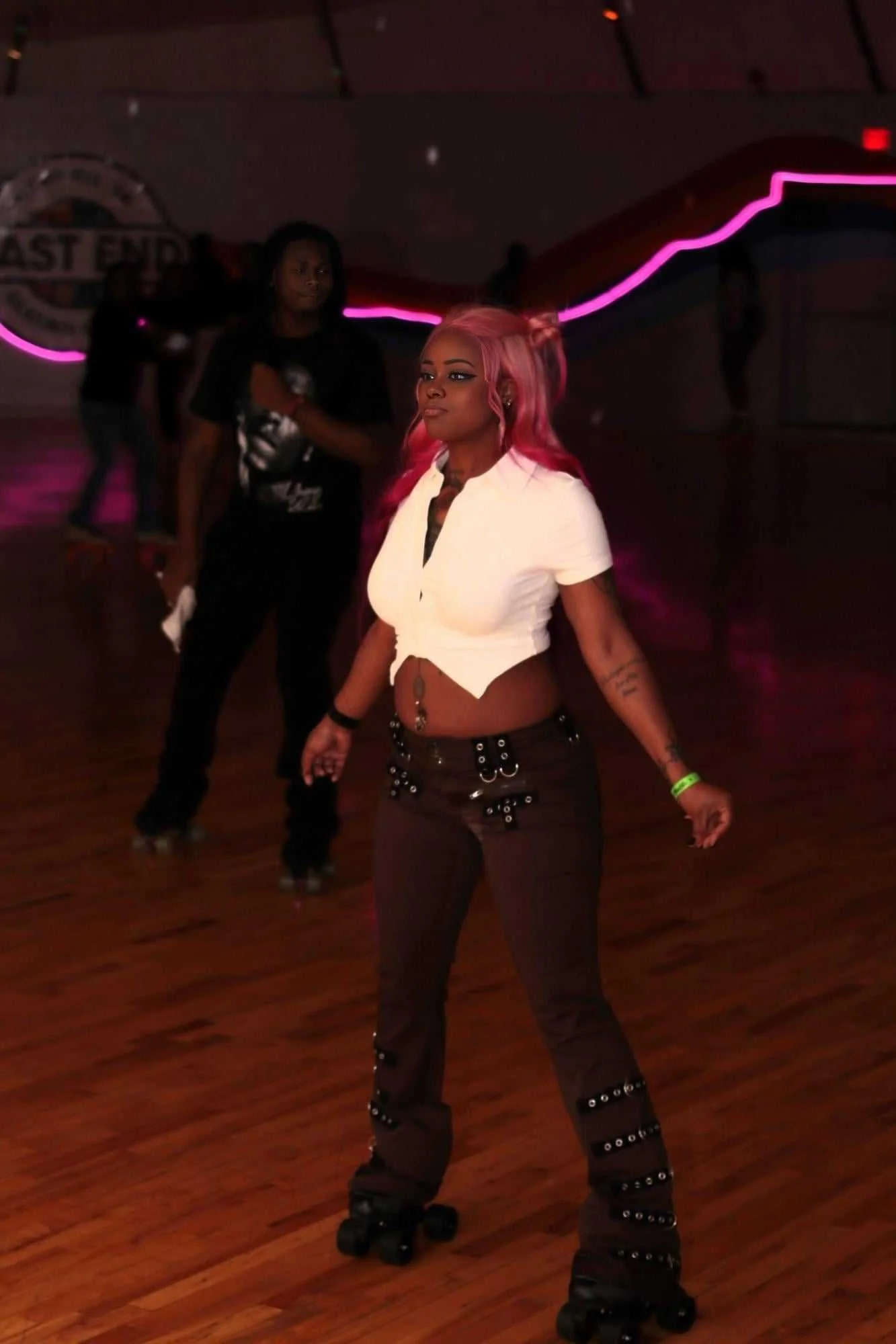 A woman with pink hair roller skating in a dimly lit indoor space with neon lights, with other people roller skating in the background.
