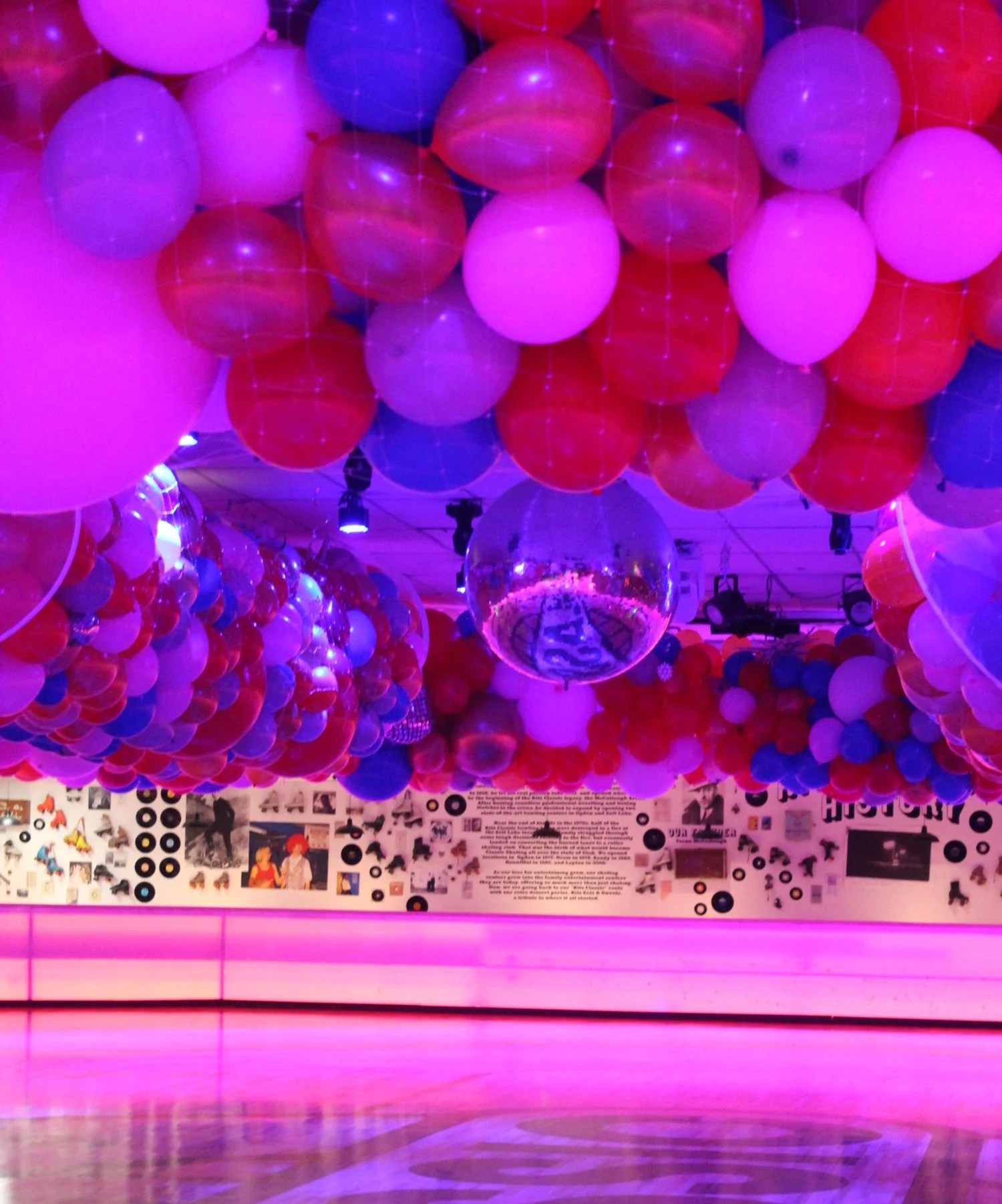 Room decorated with numerous balloons in purple, red, and white under purple lighting, with a disco ball hanging from the ceiling and a wall covered in music-themed pictures and vinyl records.