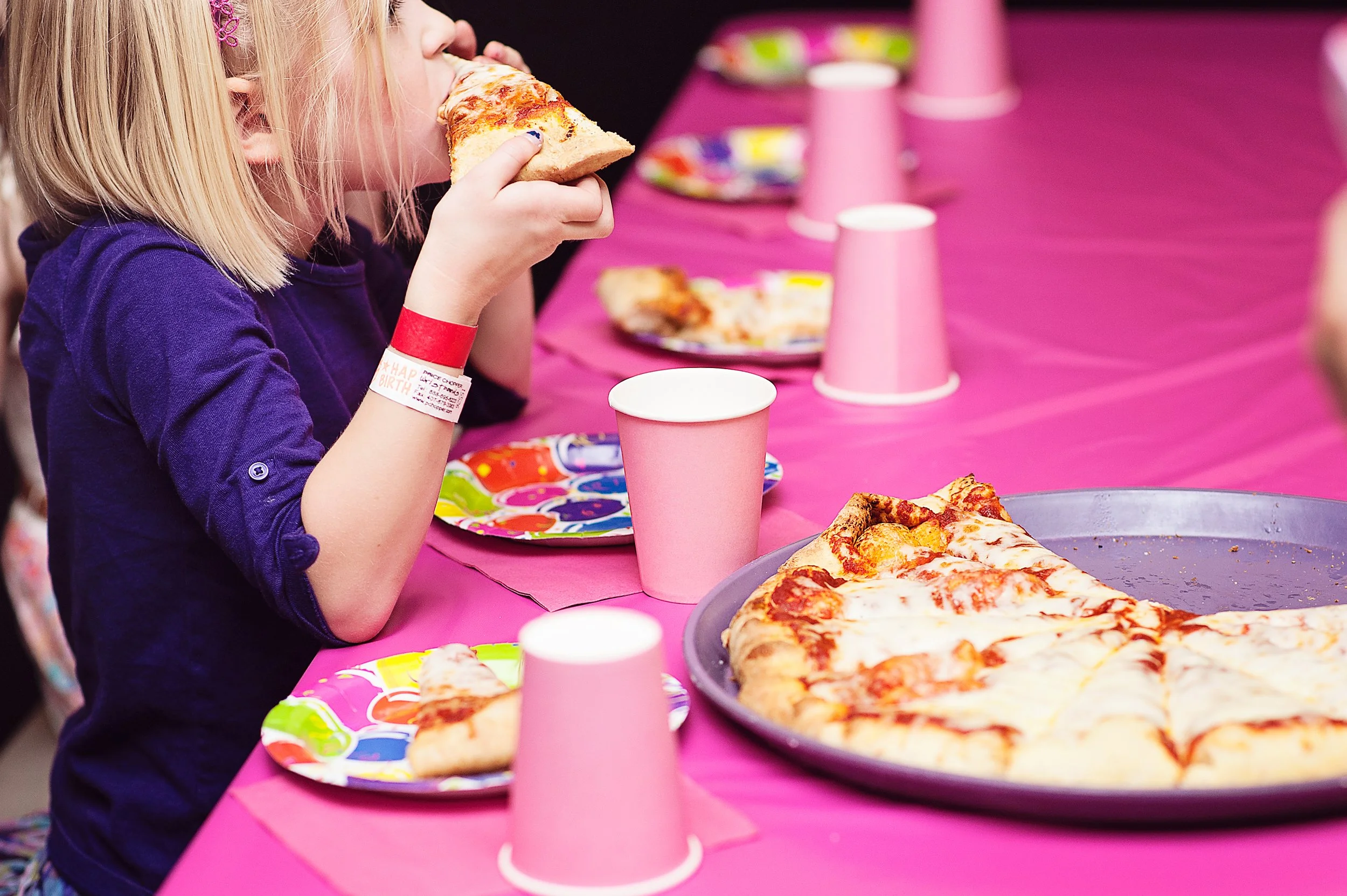 Child girl with blonde hair wearing a purple shirt at a birthday party enjoying a slice of pizza, with pink paper cups and plates on a pink tablecloth, and a large pizza on a tray in front.