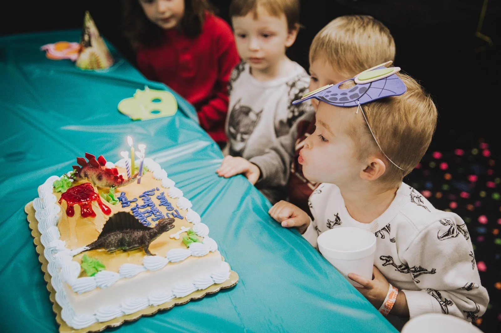 A young boy celebrating his birthday with friends, blowing out candles on a dinosaur-themed birthday cake decorated with whipped cream, fruit, and colored icing, while wearing a dinosaur hat.