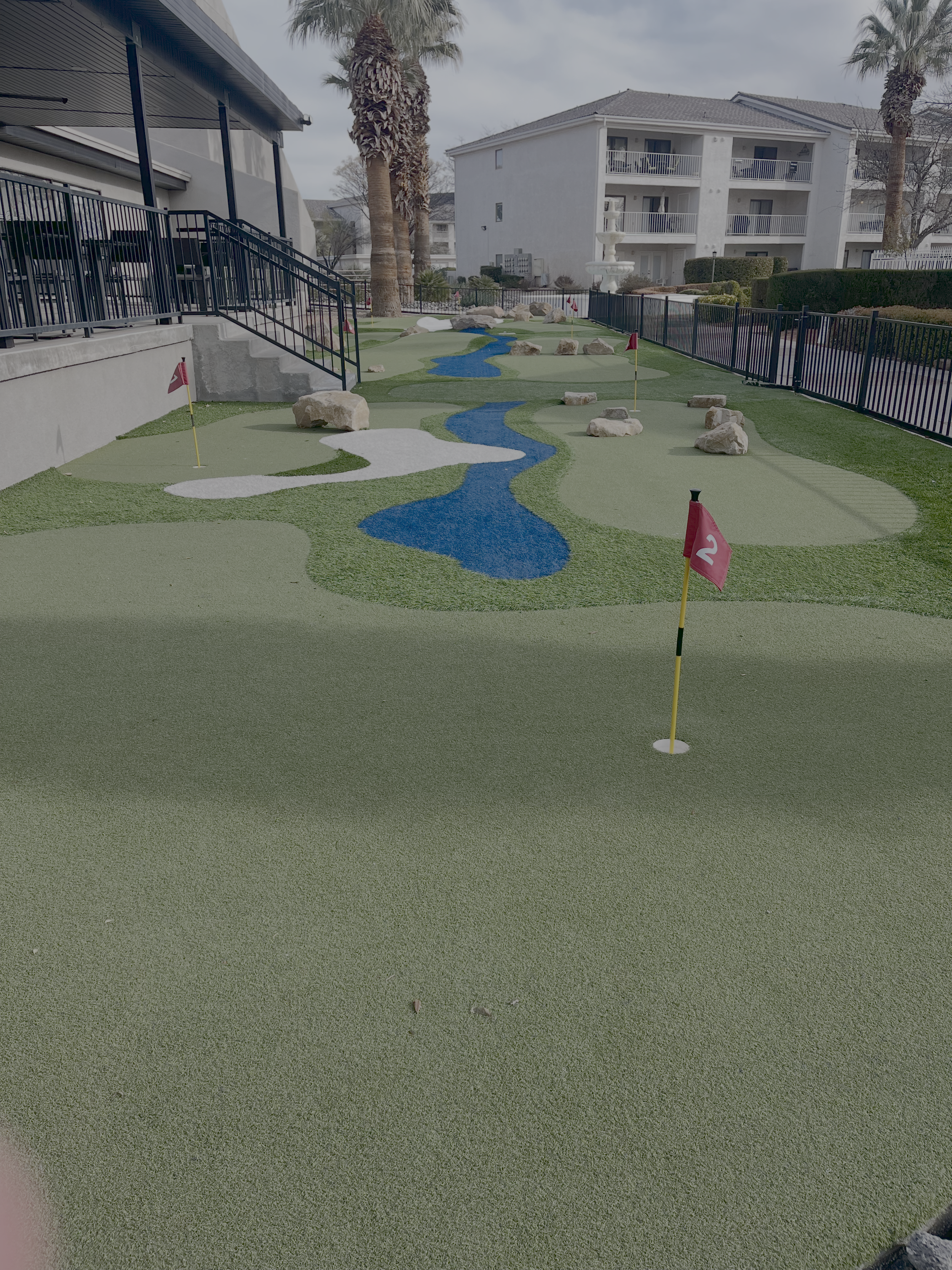 Miniature golf course with a putting green, sand traps, rocks, flags, a water feature, and surrounding buildings and palm trees.
