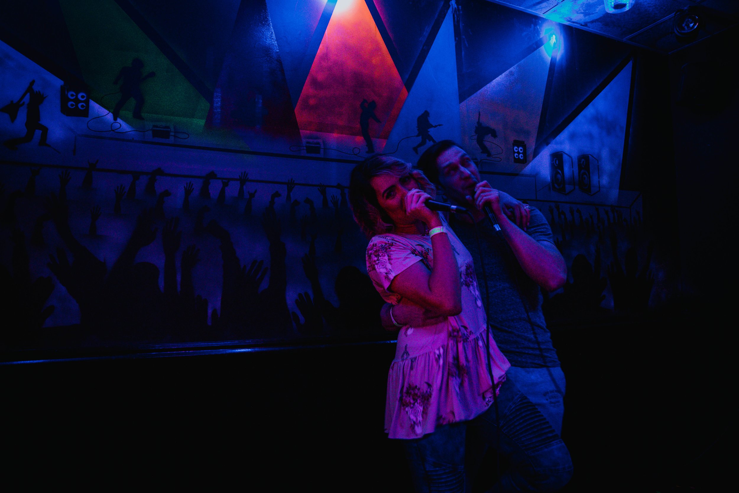 A woman and a man are singing into microphones on a stage with colorful lighting, colorful shadow silhouettes of hands and musicians in the background.