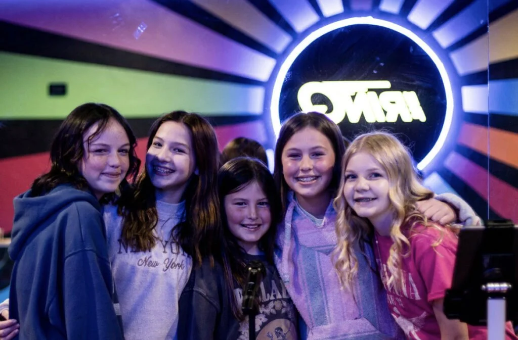 Five girls smiling and posing for a photo in front of a colorful neon sign.