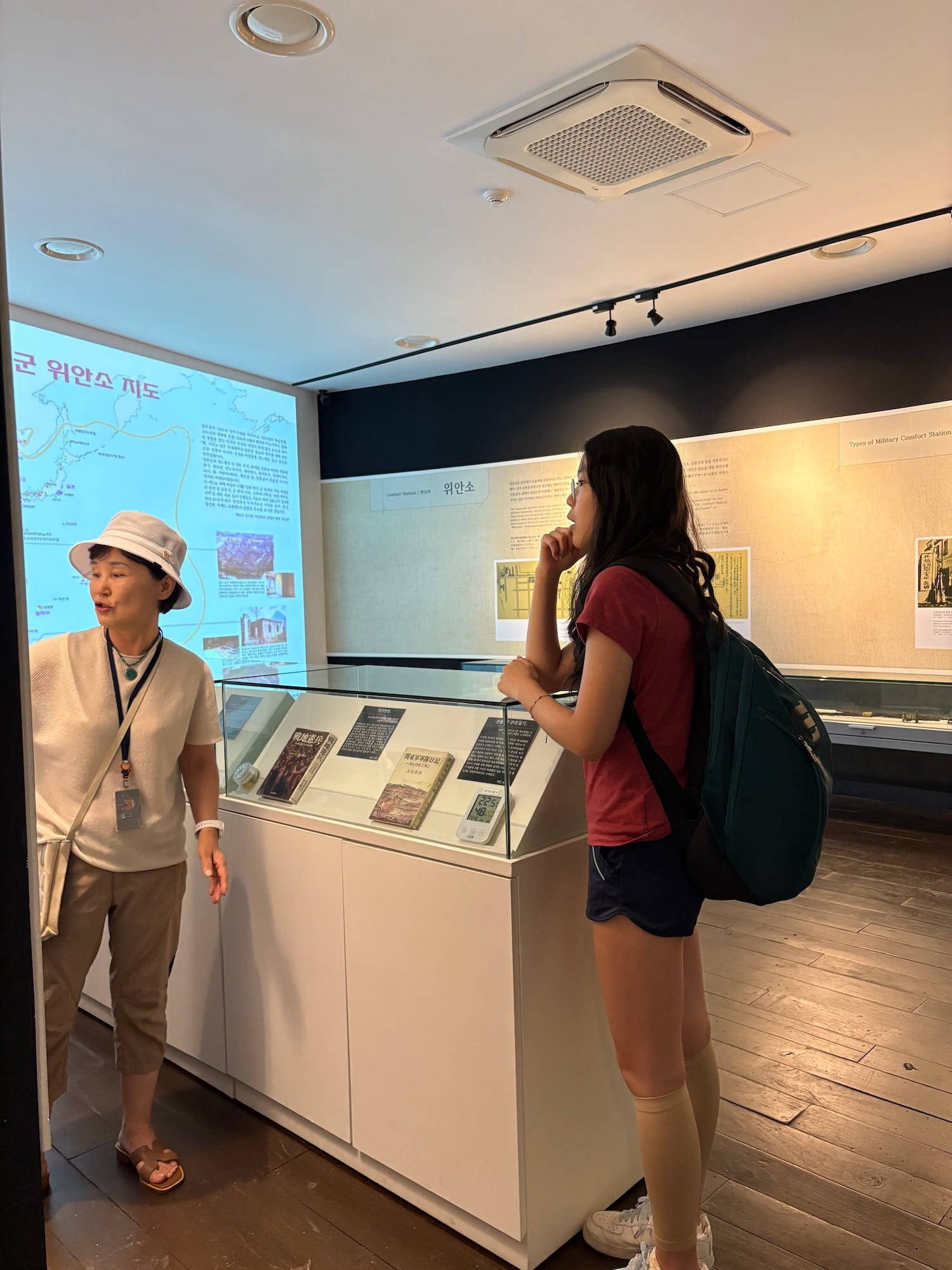 A young woman with long dark hair, wearing a red t-shirt, black shorts, and beige socks, stands in a museum, listening to a guide. The guide, an older woman with short hair, wearing a white hat, beige outfit, and sandals, explains an exhibit. Behind them are display cases with books and informational plaques, and a map projected on a screen.