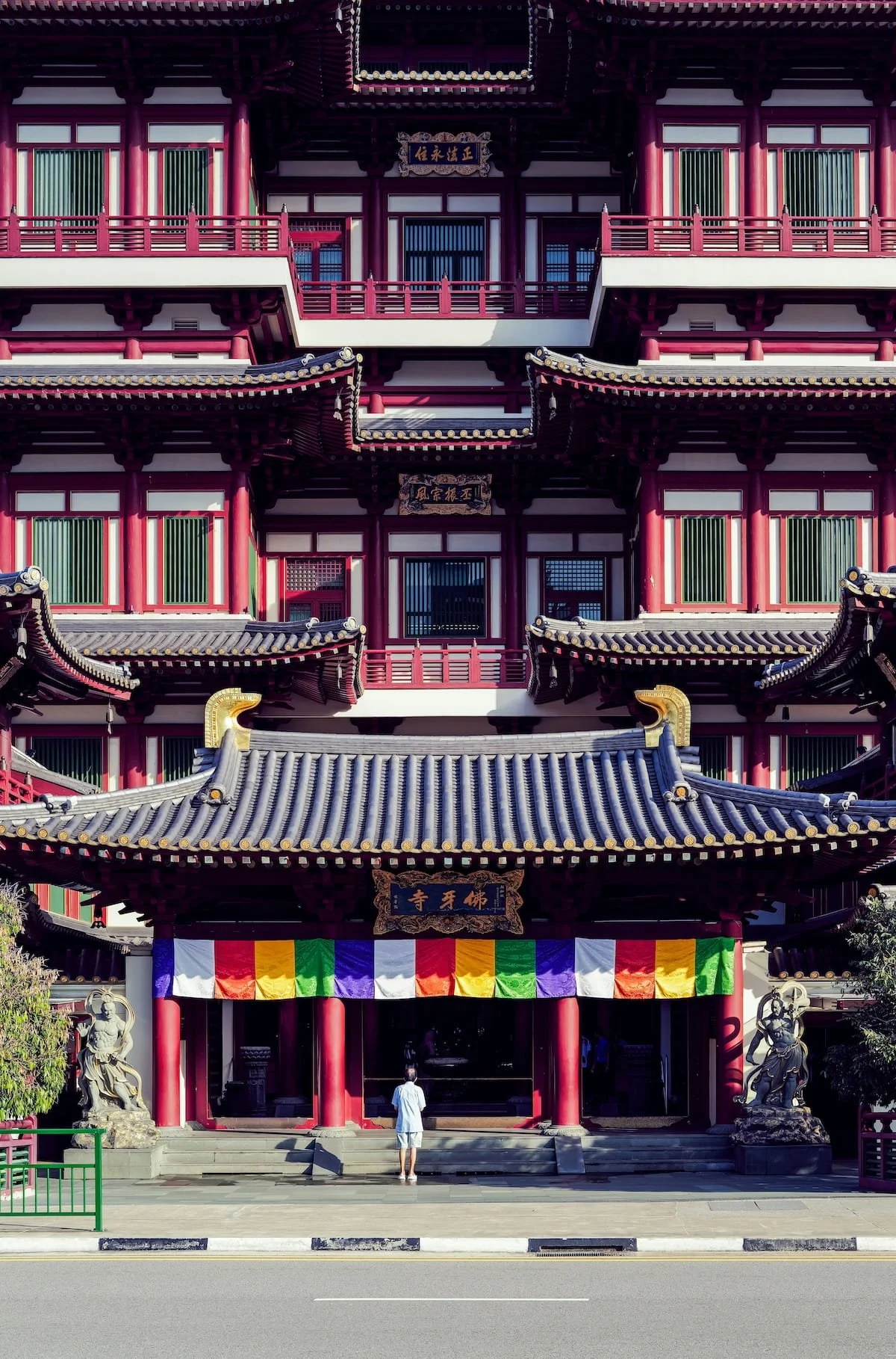 A traditional Asian temple with multiple tiers, red and black wooden architecture, curved grey-tiled roofs, colorful prayer banners, and stone statues at the entrance, with a person standing on the steps.