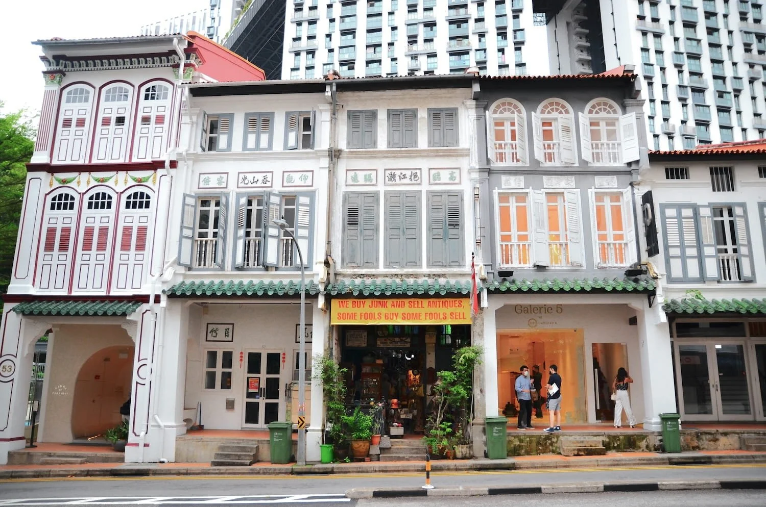 Row of colorful shophouses with traditional architecture, including shuttered windows, decorative rooflines, and signage, with people standing and talking outside, on a city street.