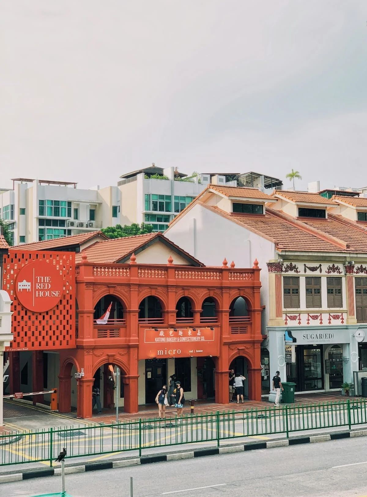 A street scene with colorful storefronts, including The Red House and Sugar D. Bakery, with people walking outside and modern buildings in the background.