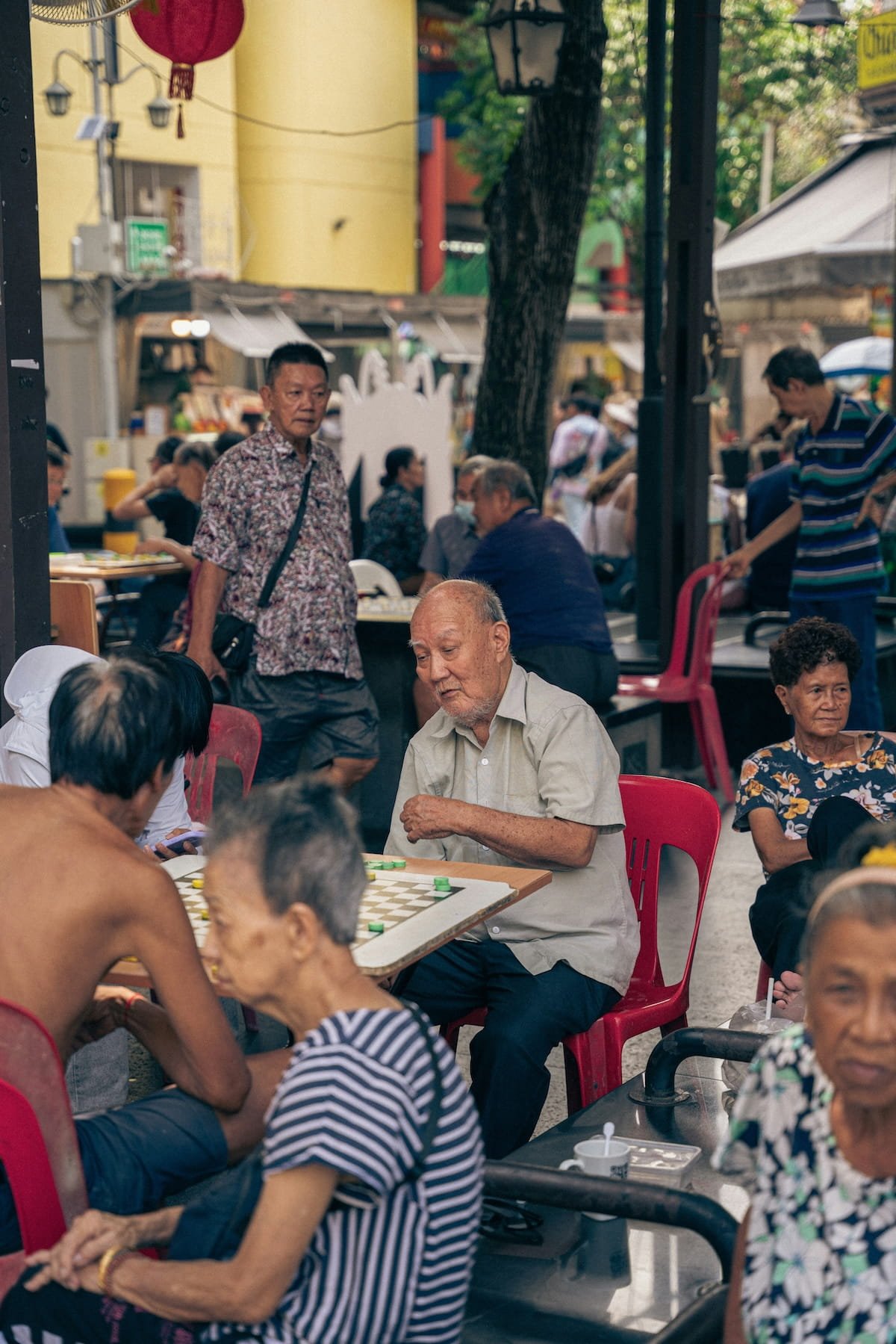 People playing checkers at an outdoor park or cafe, with several adults sitting at tables, some standing, and a street scene in the background.