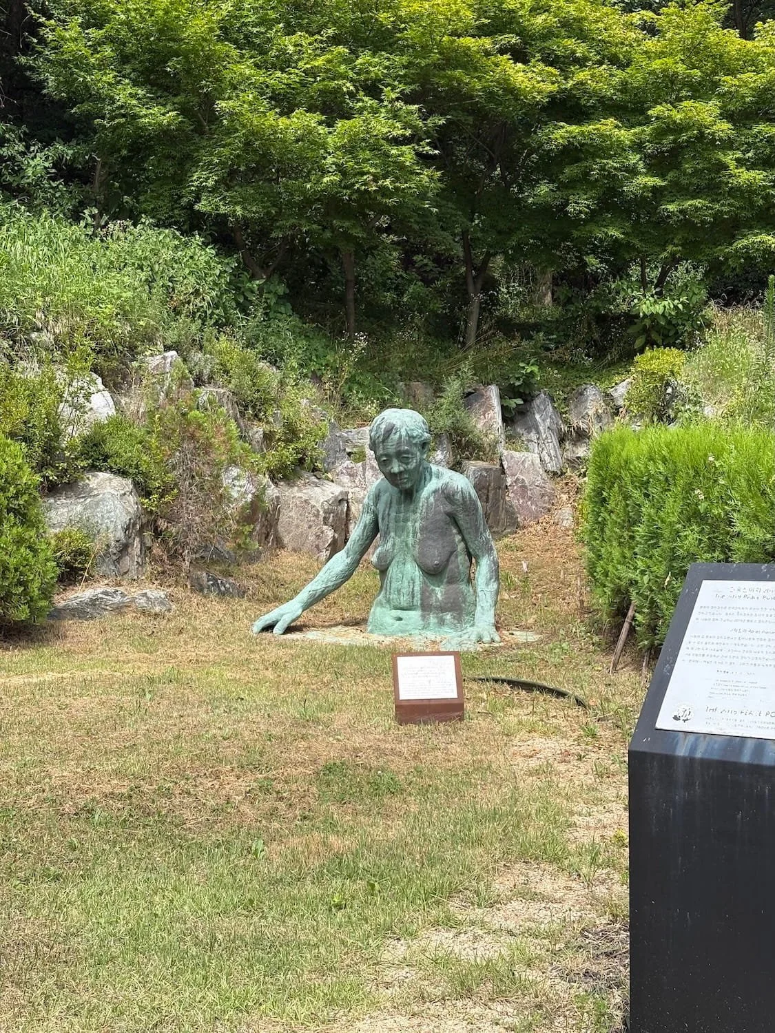 Outdoor sculpture of a woman with missing upper torso, emerging from the ground, with trees and bushes in the background.