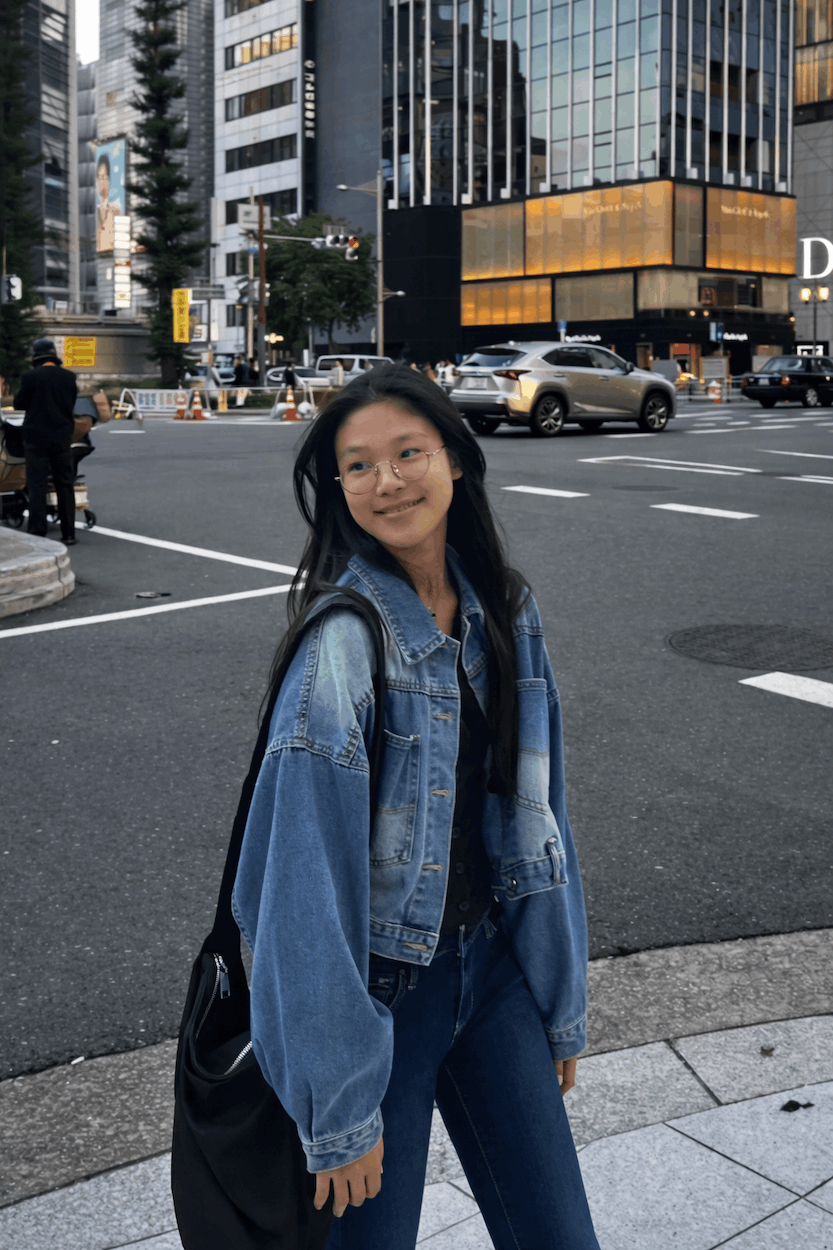 A young woman with glasses and long black hair wearing a blue denim jacket, standing on a city street with tall buildings, cars, and pedestrians in the background.