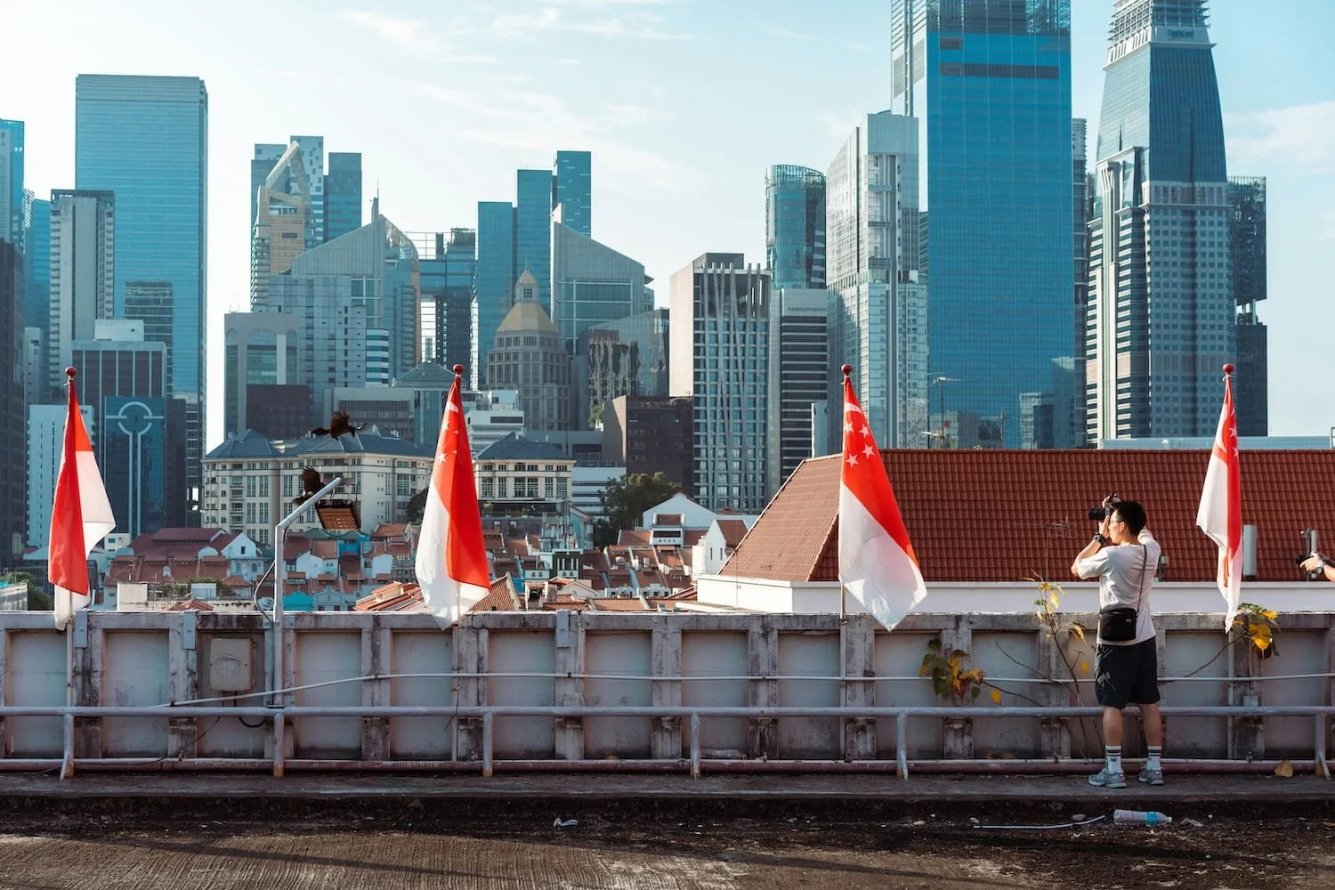 A man shooting a camera on a rooftop with Indonesian flags in the foreground and a city skyline with modern skyscrapers in the background.