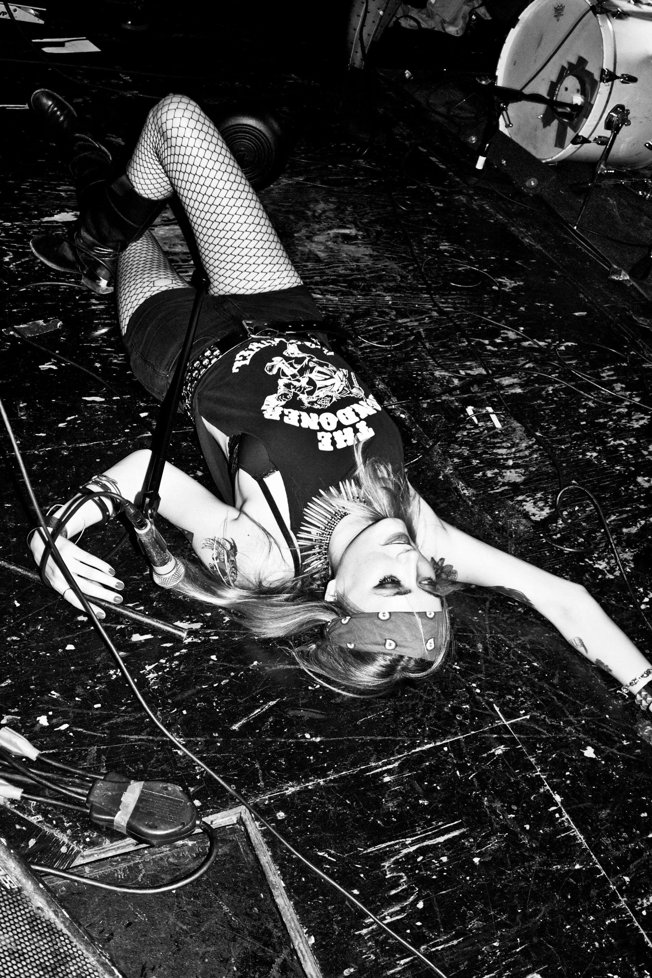A woman lying on the stage floor with her eyes closed, holding a microphone, dressed in punk style clothing with fishnet stockings and a bandana, near musical instruments.