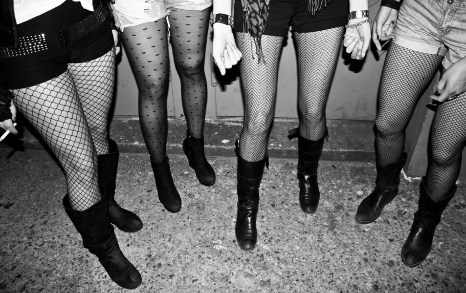Group of women wearing fishnet tights and boots, holding hands, standing on concrete floor.