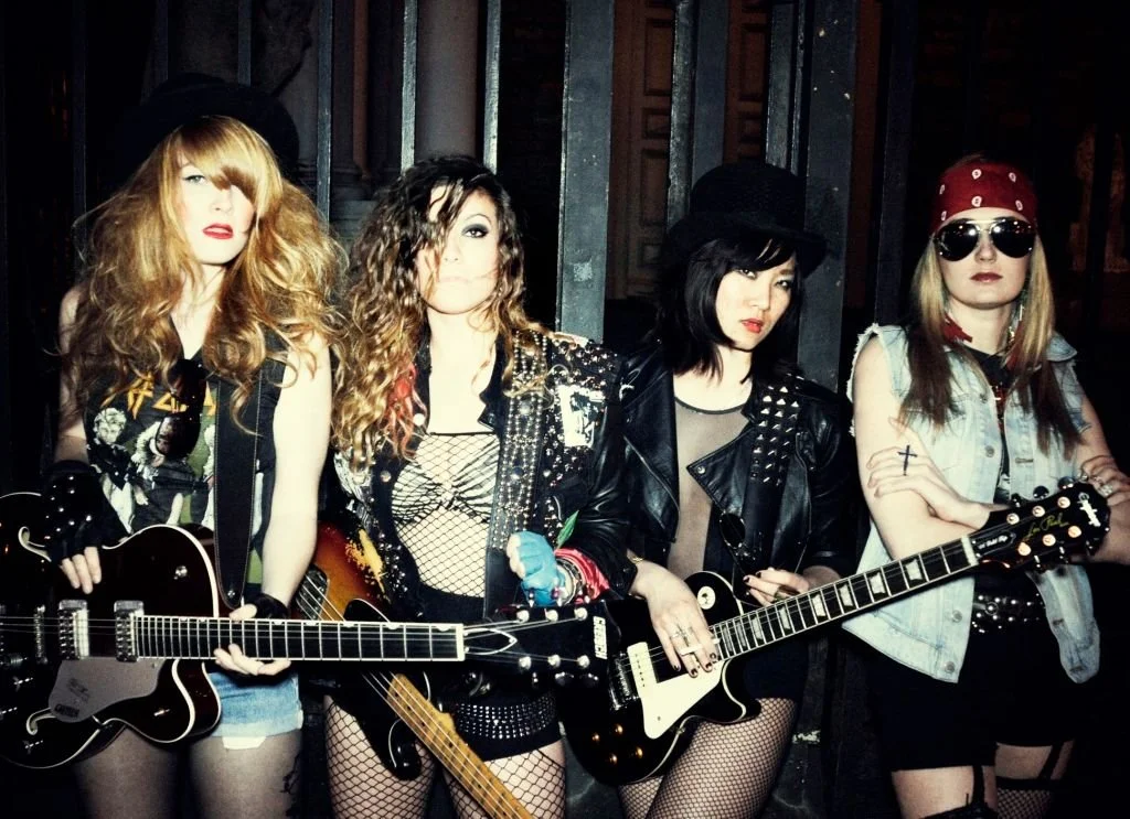 Four women dressed punk rock style holding electric guitars, with a dark background.