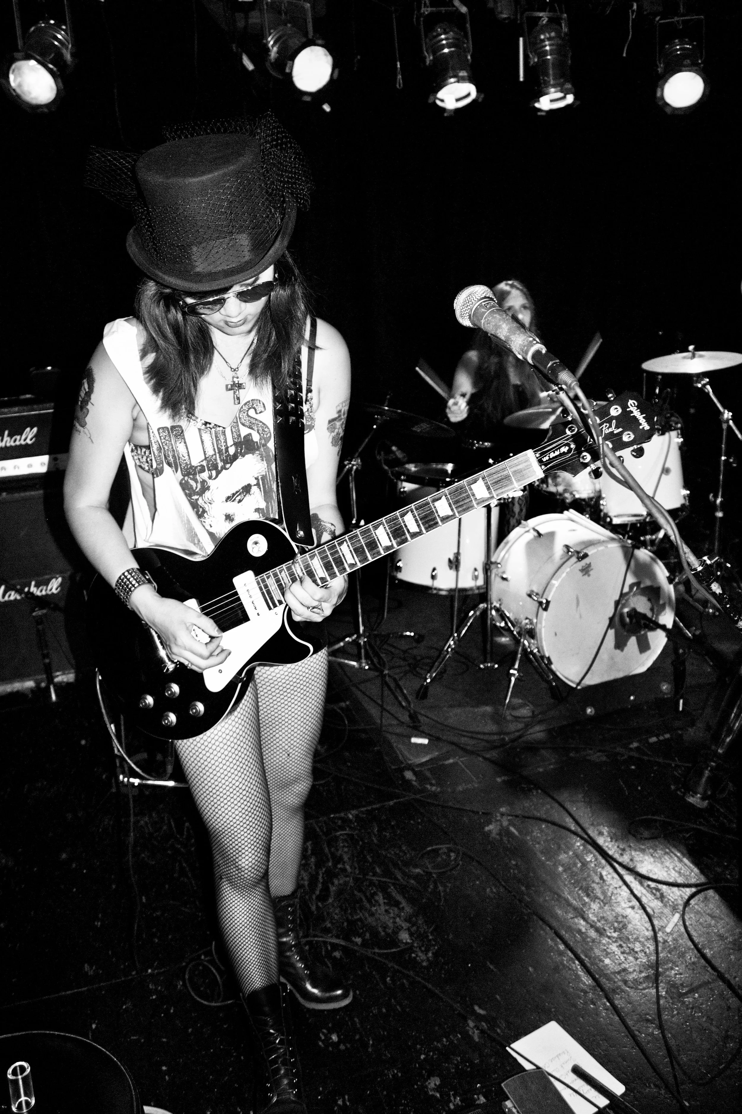 A woman wearing sunglasses, a hat, fishnet stockings, and a Rolling Stones T-shirt plays electric guitar on stage with drums and amplifiers behind her.