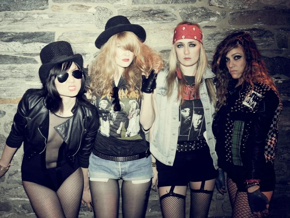 Four women dressed in punk and rock-inspired outfits, standing against a stone wall, with edgy makeup, fishnet stockings, and leather accessories.