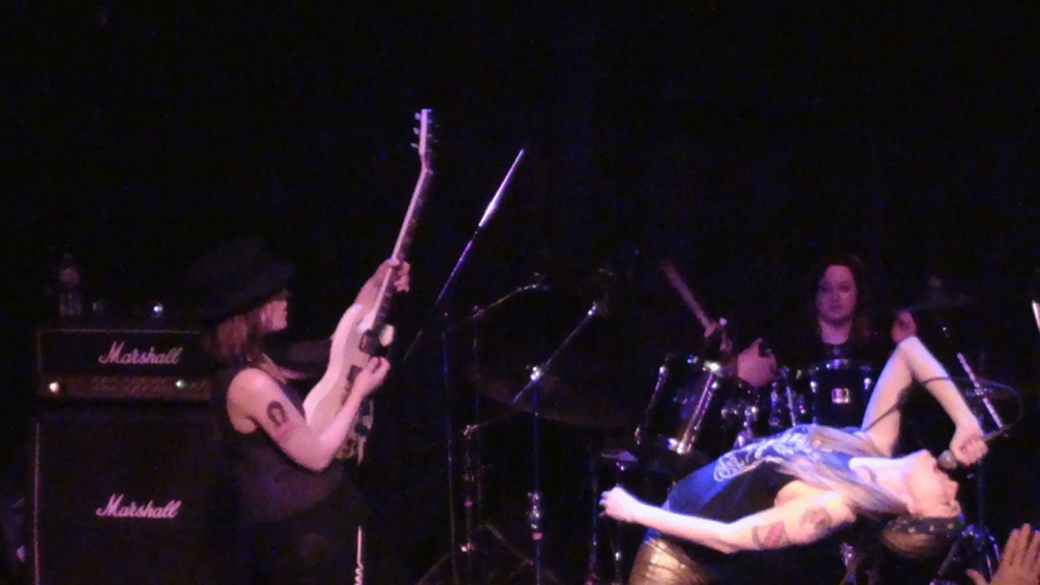 Two female musicians on stage, one playing an electric guitar and the other on drums, with a dark background and stage lighting.