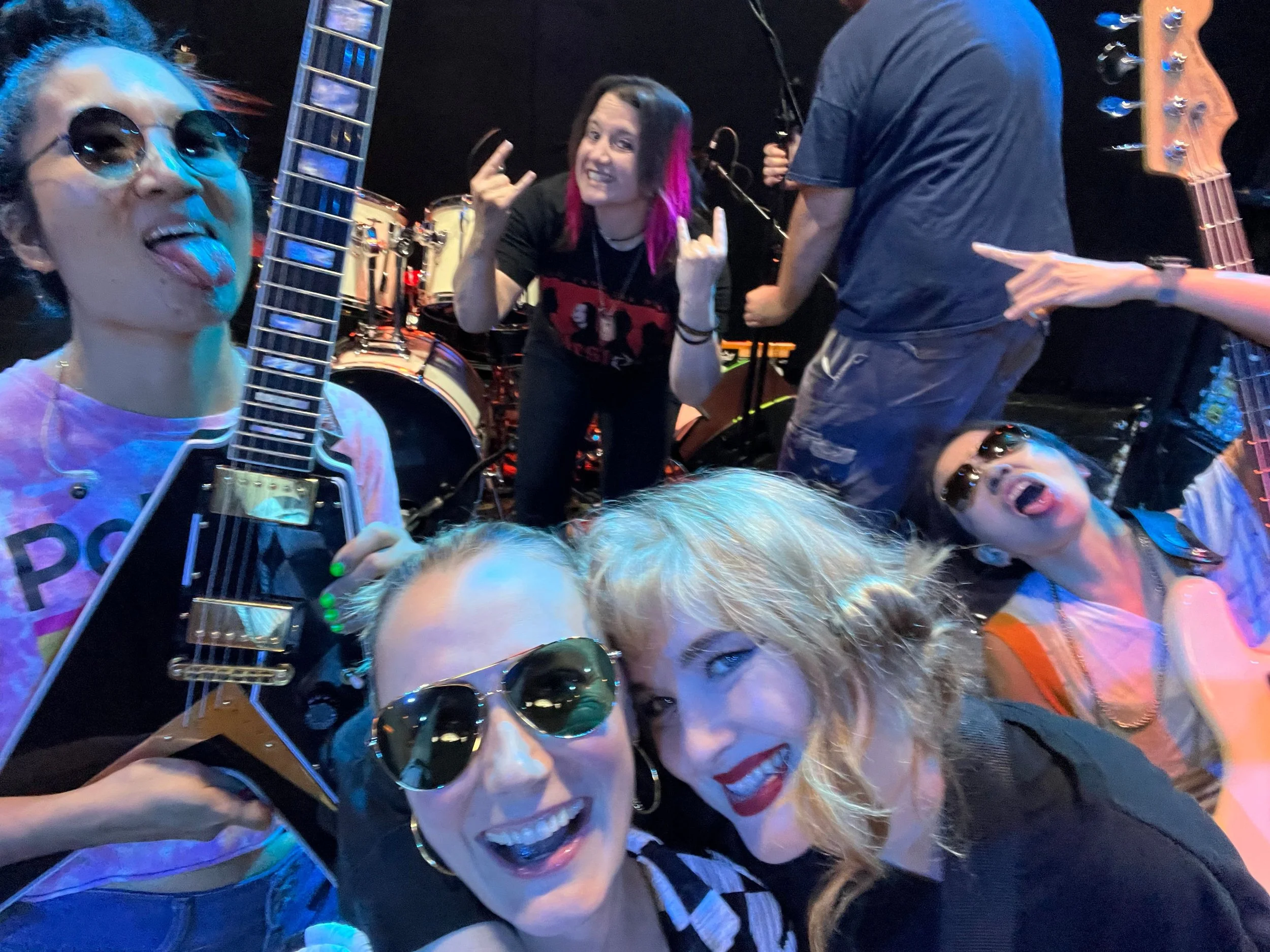 Group of five women at a music venue, one holding a guitar, with drums and a bass guitar in the background, all wearing sunglasses and expressing lively emotions.