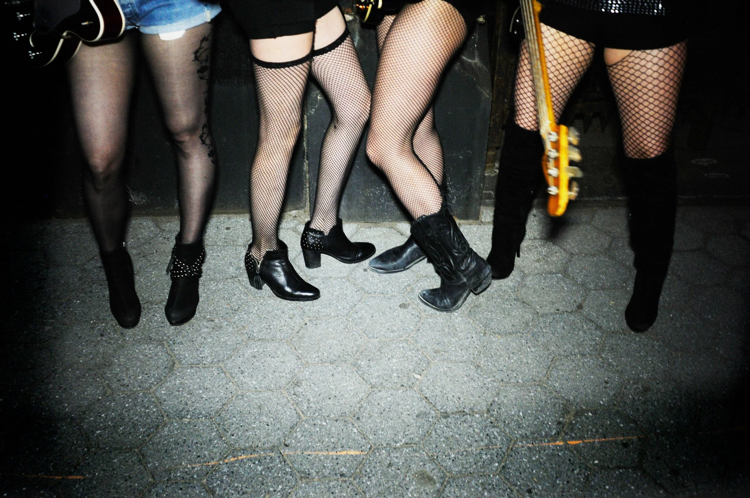 Close-up of four women standing together wearing black fishnet stockings and black footwear, with a guitar visible in the shot, on a textured outdoor surface at night.