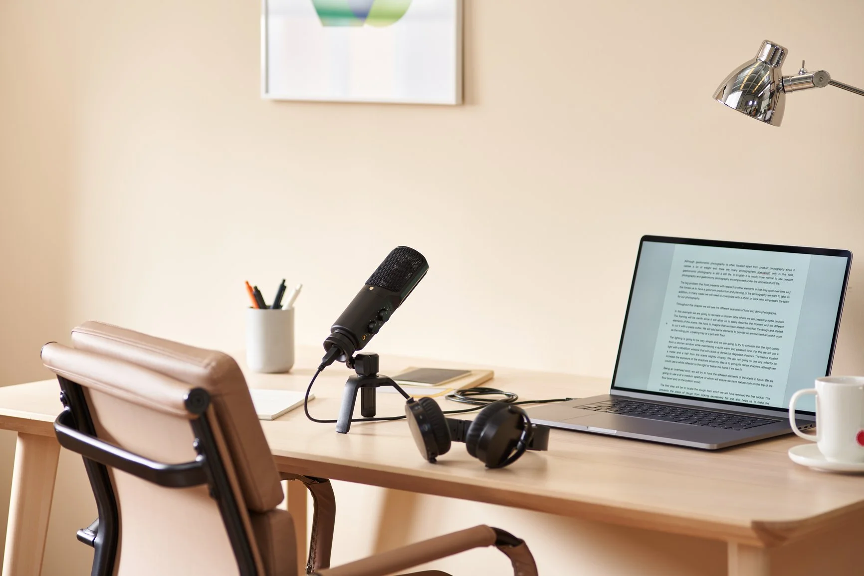 A tidy home office desk with a laptop displaying text in a document, a black microphone on a stand, closed black over-ear headphones, a beige office chair, a white mug, a container of pens, and a desk lamp, all against a beige wall with a framed picture.