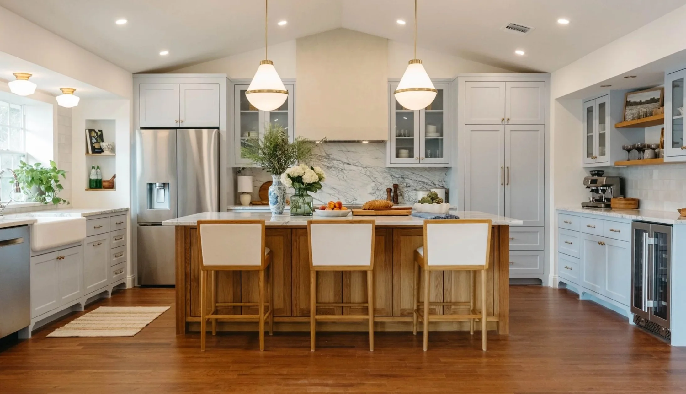 A spacious kitchen with white cabinets, a central island with a wooden base, and modern appliances. There are two pendant lights hanging above the island, a vase with flowers, and a variety of kitchen items such as bread, apples, and decorative piece