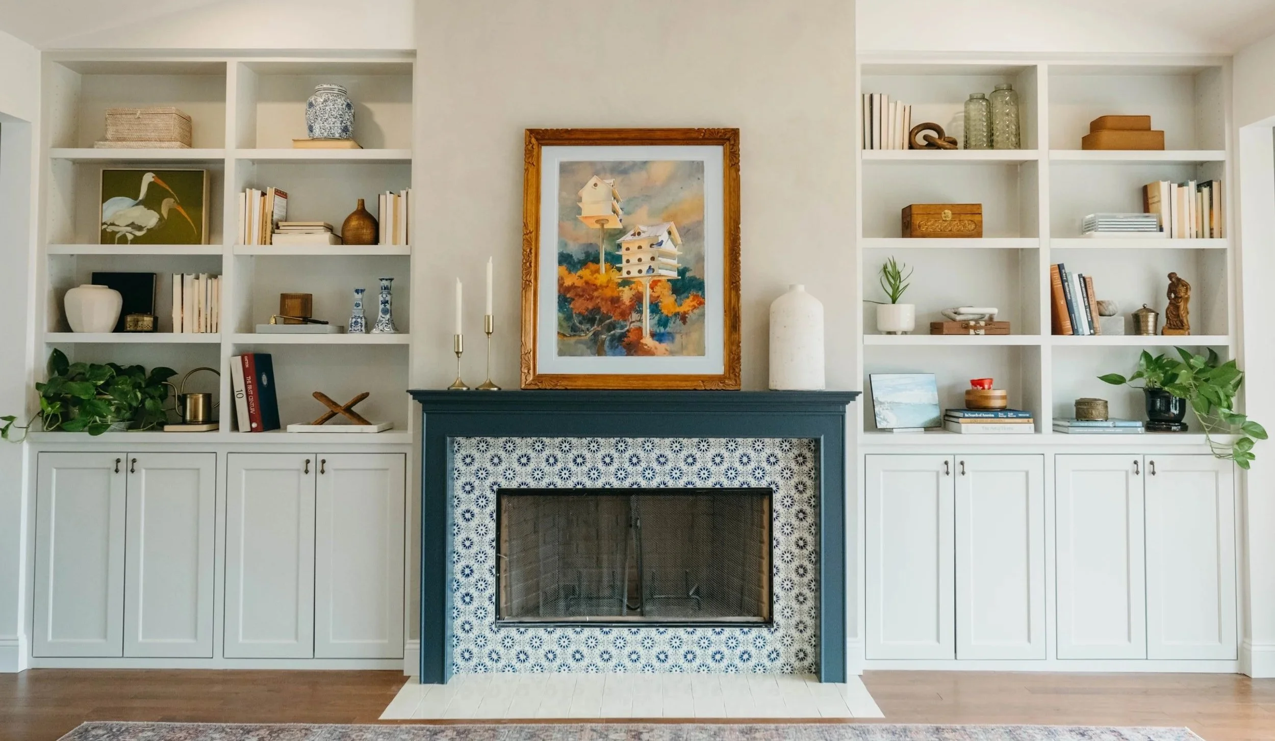 A living room fireplace with a blue mantle, surrounded by built-in white bookshelves filled with books, decorative items, and plants. A framed picture of birdhouses hangs above the fireplace.