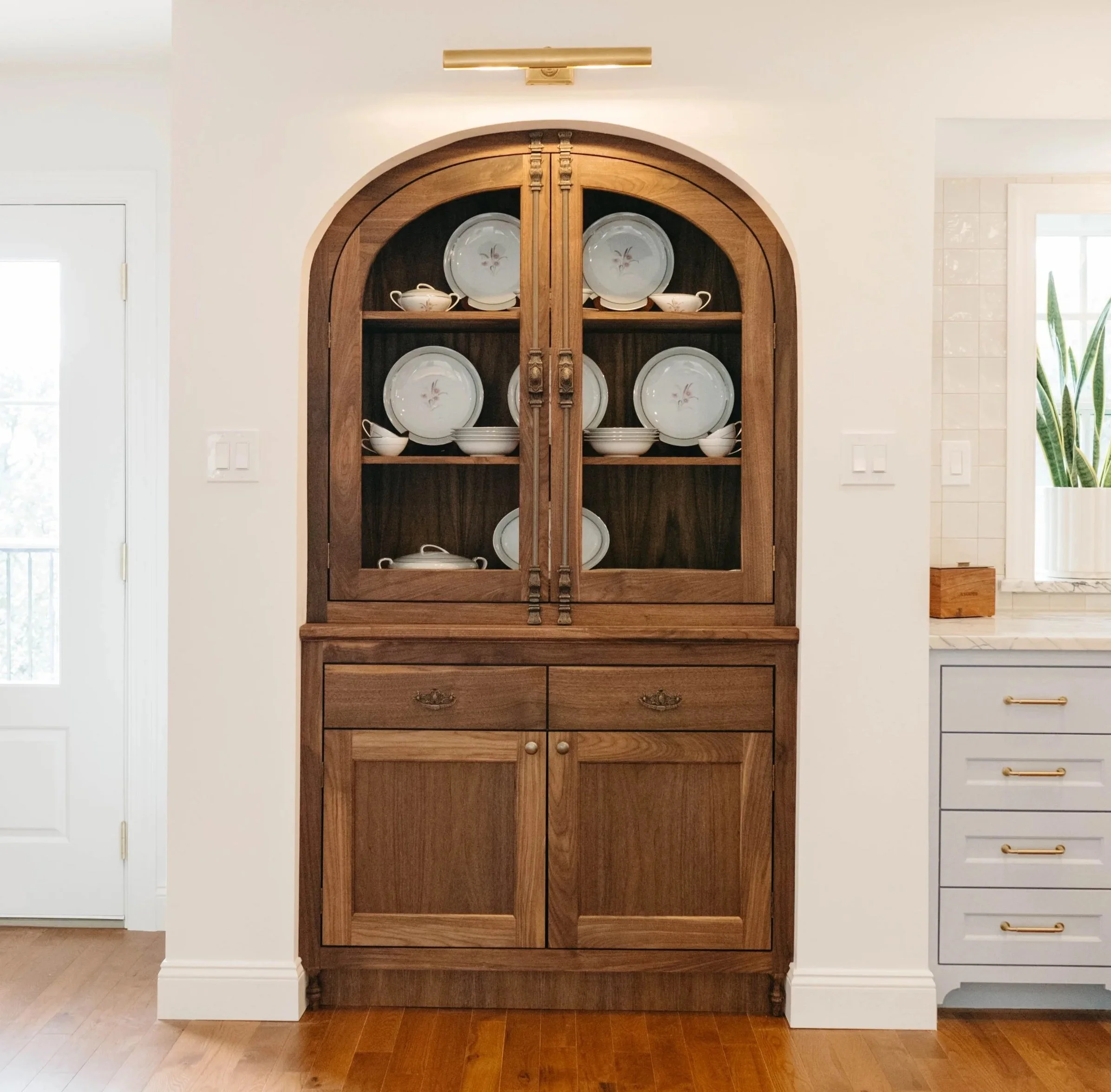 A custom walnut china cabinet display cabinet with glass doors containing white dishware with floral designs, set against a white wall in a bright room.