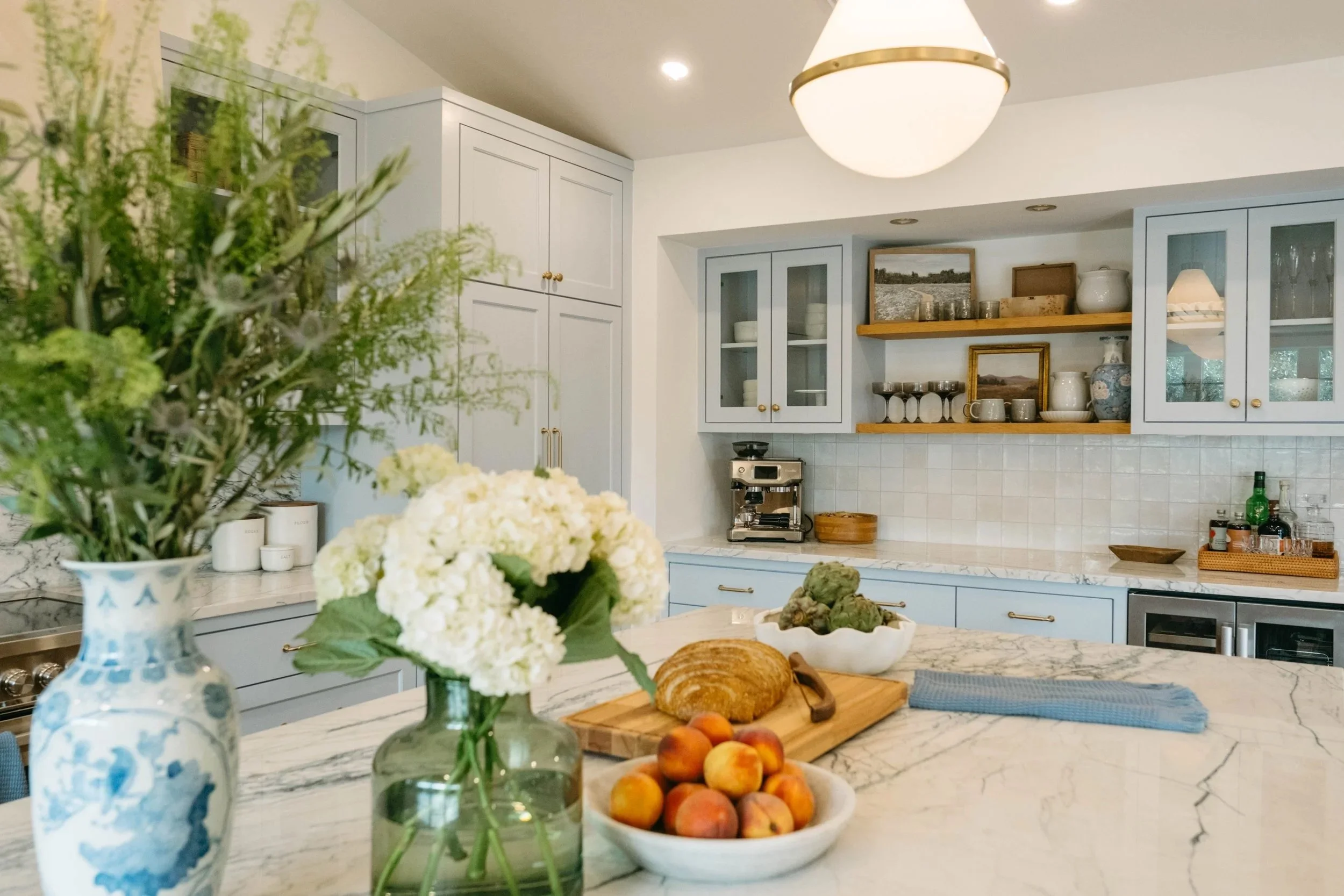 A kitchen with light blue cabinets and open shelves, decorated with various dishes and decor items. A vase with green foliage and white flowers is in the foreground. The countertop has a loaf of bread, peaches, artichokes, and a blue cloth. The ceiling has a large round light fixture, and the kitchen has white tiled walls.