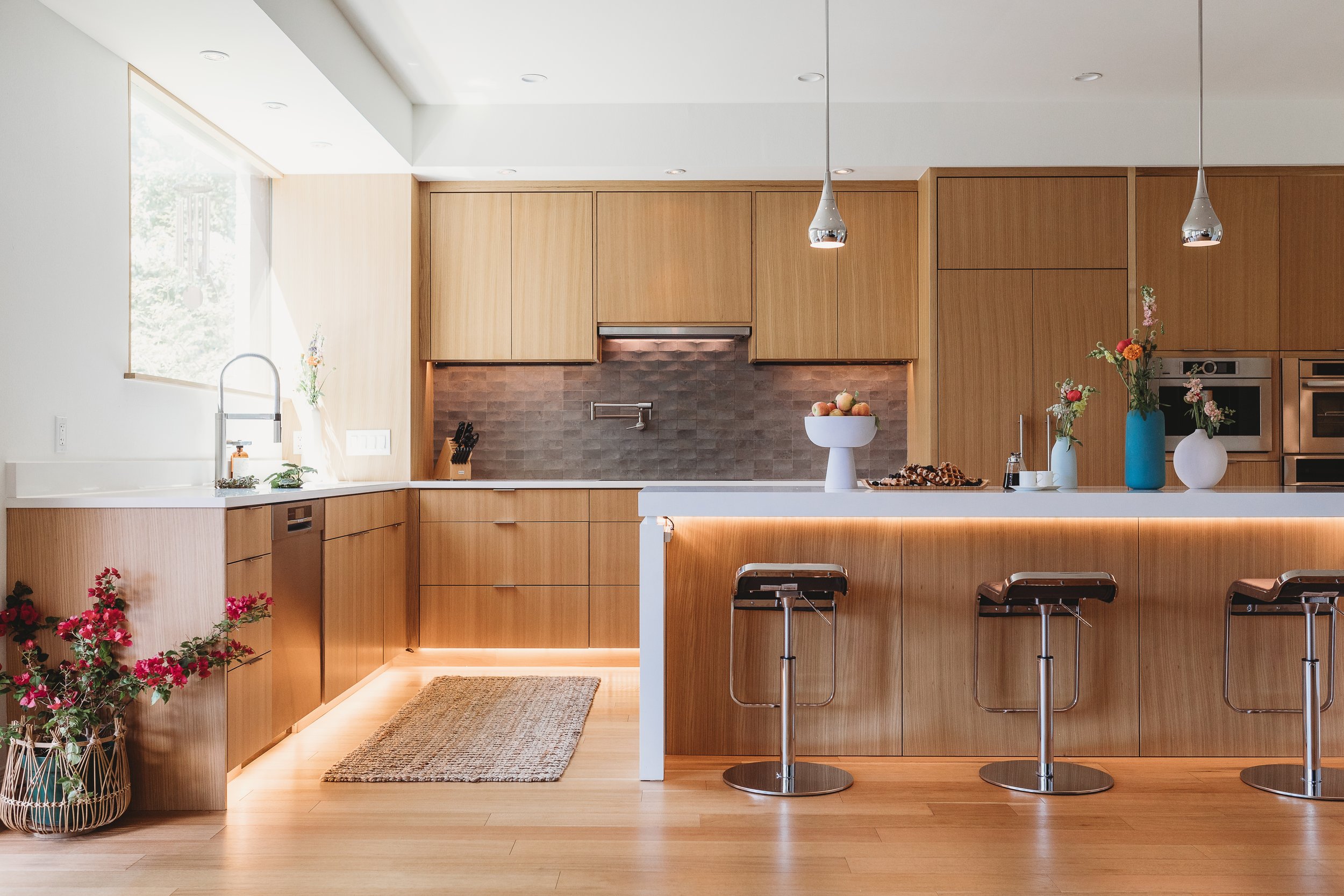 Modern kitchen with custom wood cabinets, white countertops, chrome pendant lights, floral vases, and a fruit bowl on the island.