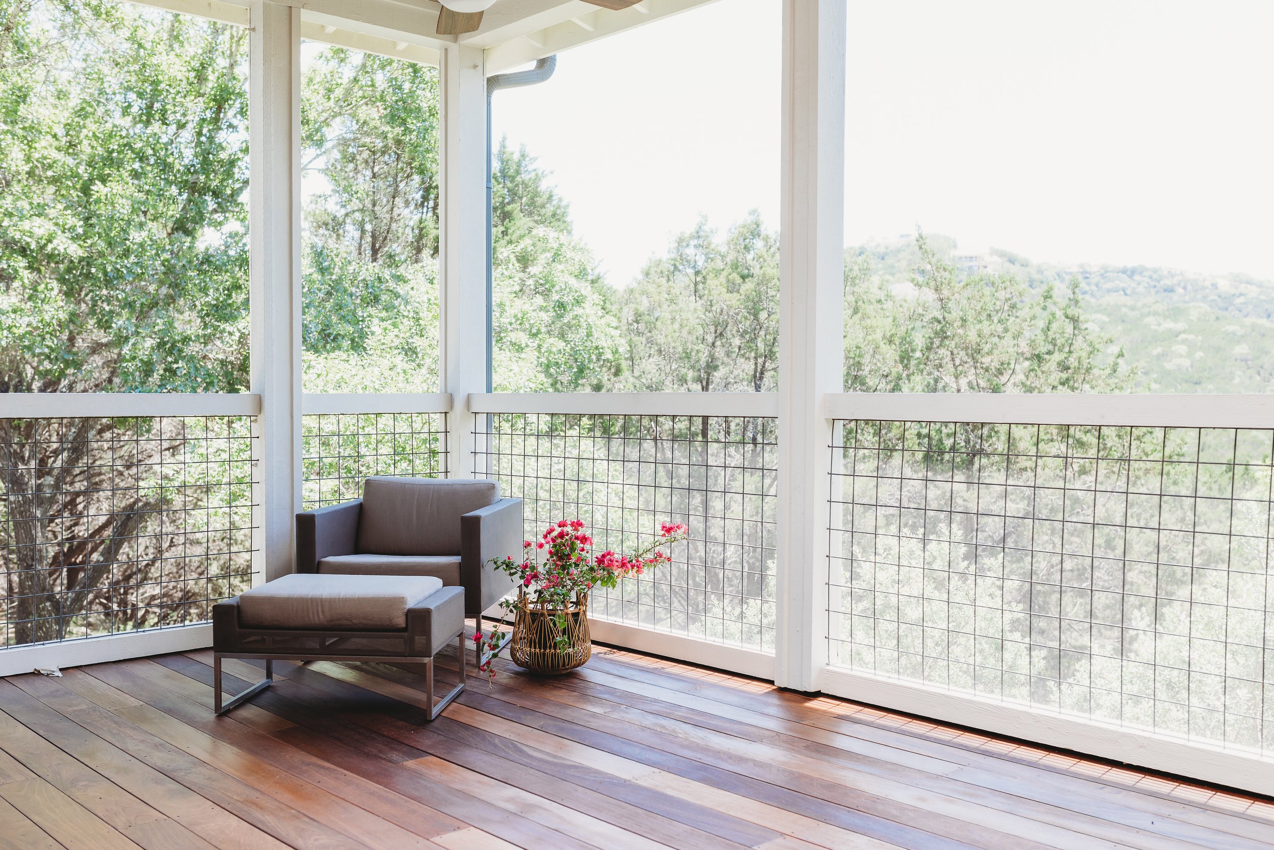 A screened porch overlooking trees and greenery, furnished with a small beige armchair, a boxy ottoman, and a decorative basket with pink flowers, illuminated by bright sunlight.