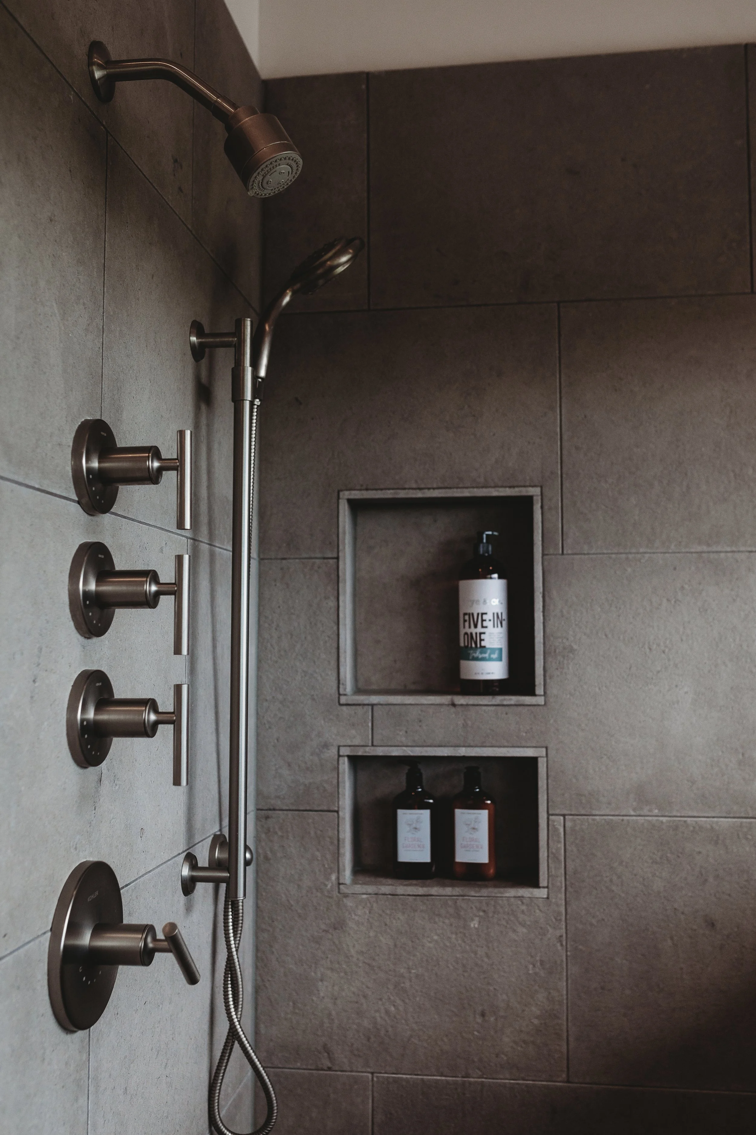 A modern bathroom shower with a rain shower head, multiple control knobs, and built-in wall niches with bottles of soap and shampoo.