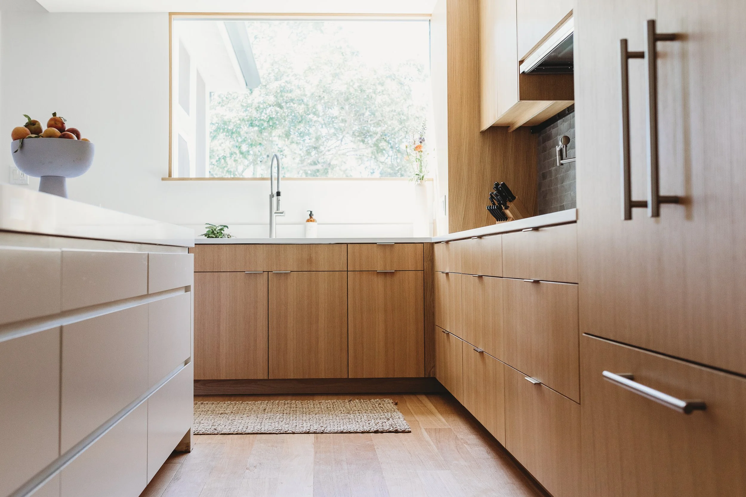 Kitchen with wooden cabinets, a large window, a sink, a bowl of fruit on a white counter, and a kitchen knife block on the counter.