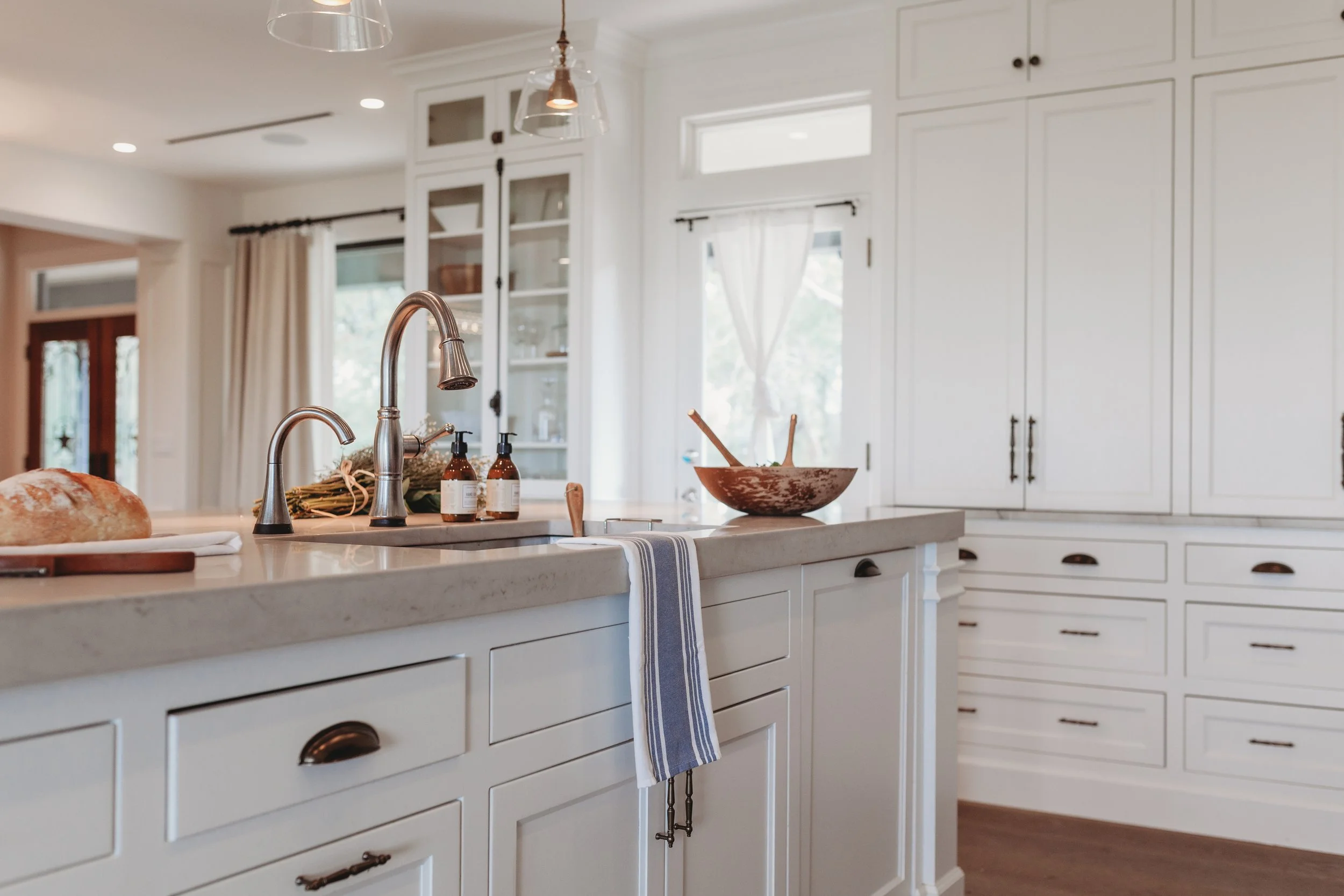Modern white kitchen with marble countertop, bronze sink fixtures, glass-front cabinets, and a wooden bowl with cooking utensils