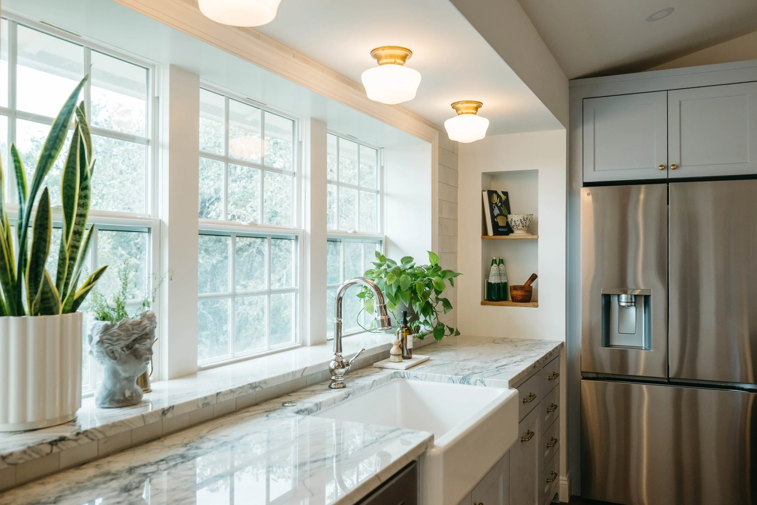 Bright kitchen with large windows, marble countertop, potted plants, decorative bust, and stainless steel refrigerator with a water dispenser.