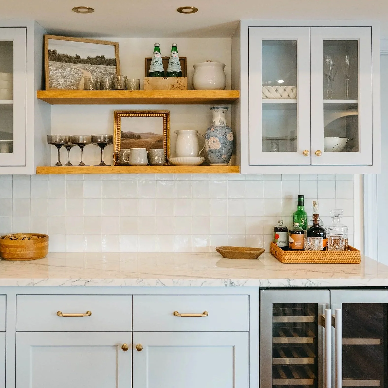 White kitchen cabinetry with glass front upper cabinets, open wooden shelves with glassware and framed landscape photos, marble countertop, and a wine fridge.