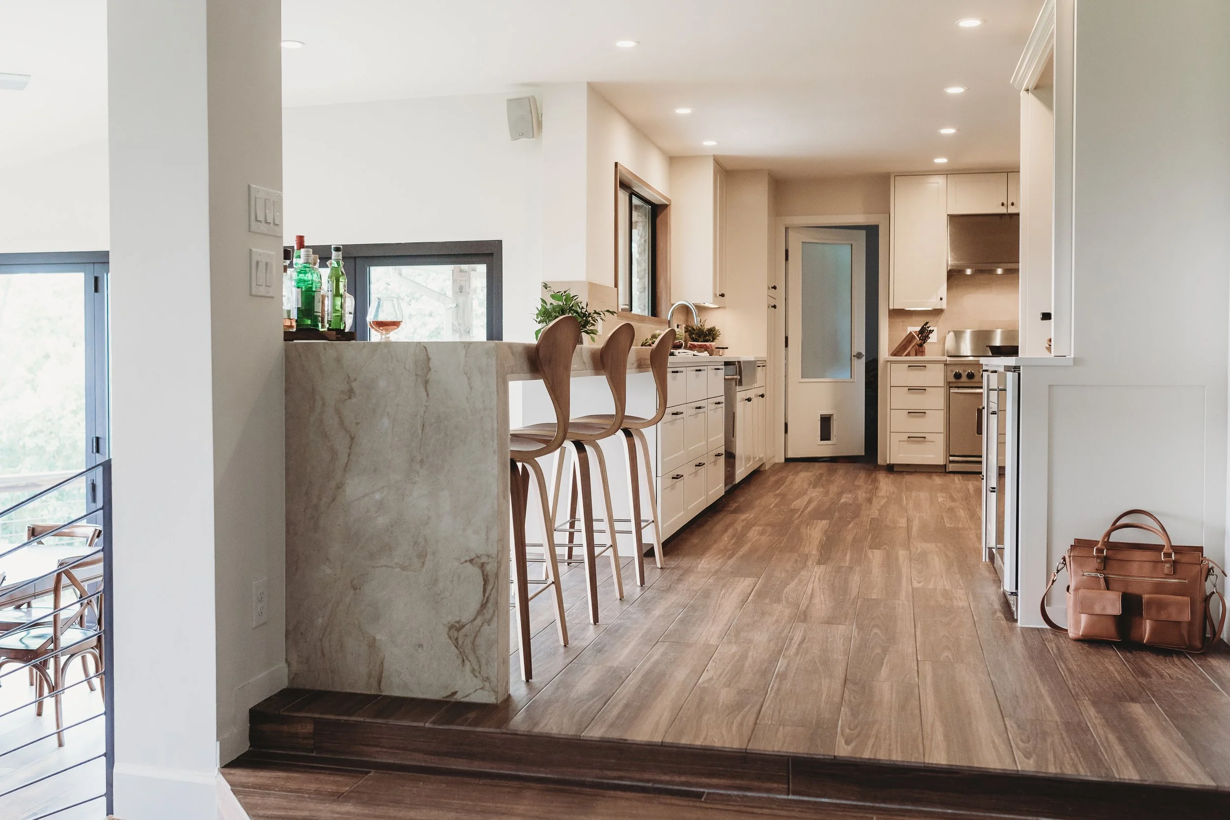 Interior view of a modern kitchen with wooden flooring, white cabinets, a marble kitchen island with barstools, and large windows.