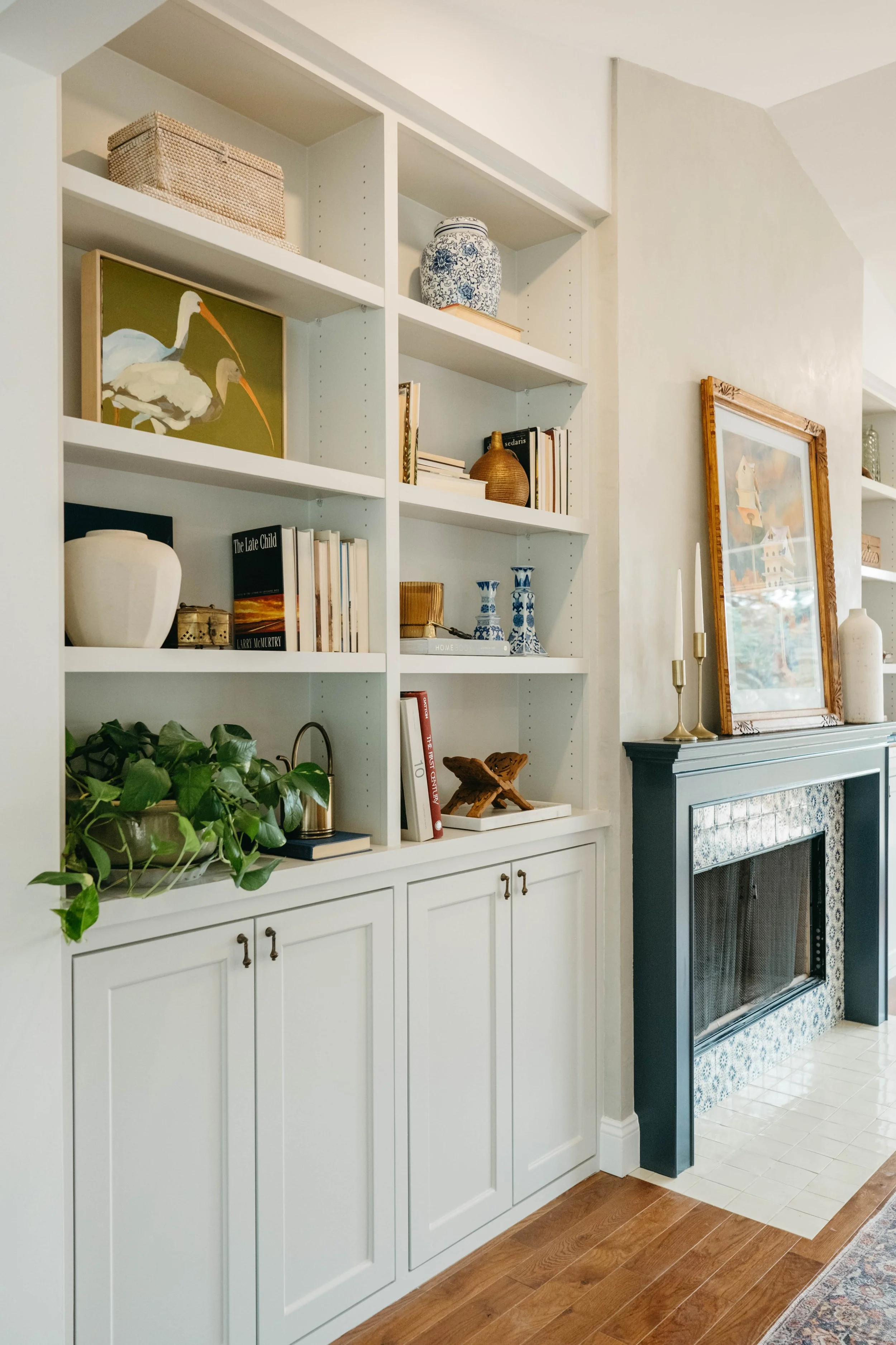 A white built-in bookshelf filled with books, decorative vases, and artwork next to a fireplace with a framed painting and brass candlesticks.
