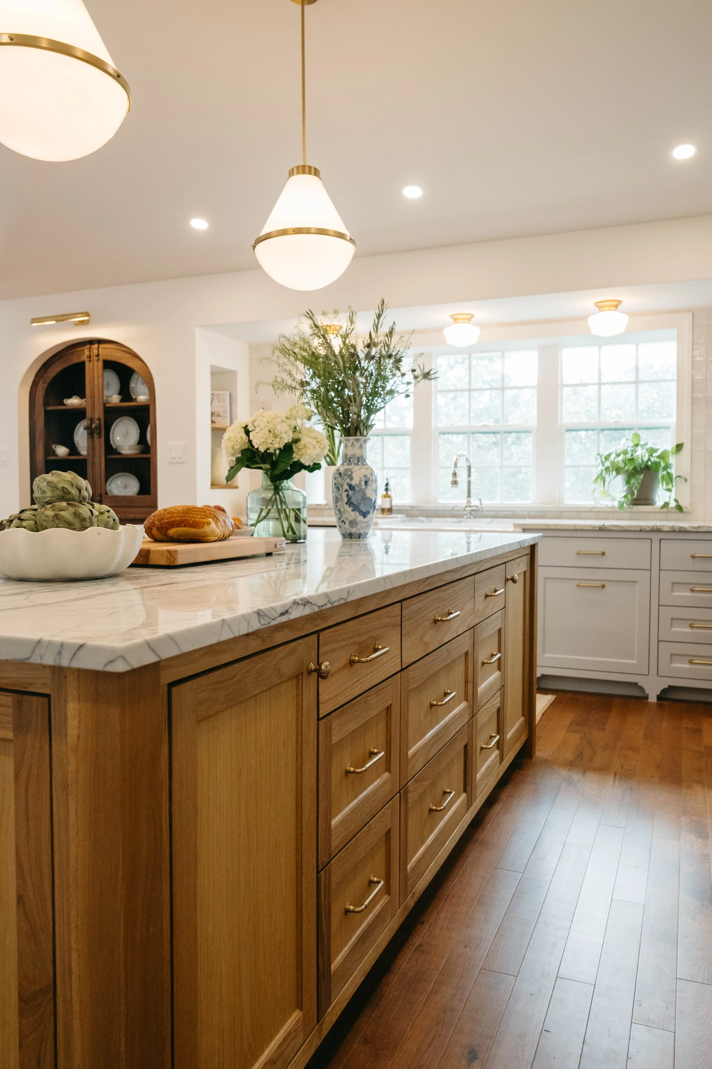 A kitchen with a wooden island, marble countertop, and white cabinets. Decor includes vases with flowers, a bowl of artichokes, and a loaf of bread. Large windows allow natural light, and ceiling lights illuminate the space.